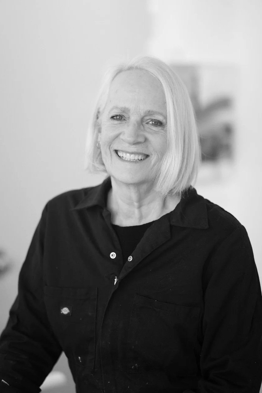 A black-and-white photograph of an older woman with shoulder-length blonde hair, smiling and wearing a dark button-up shirt.