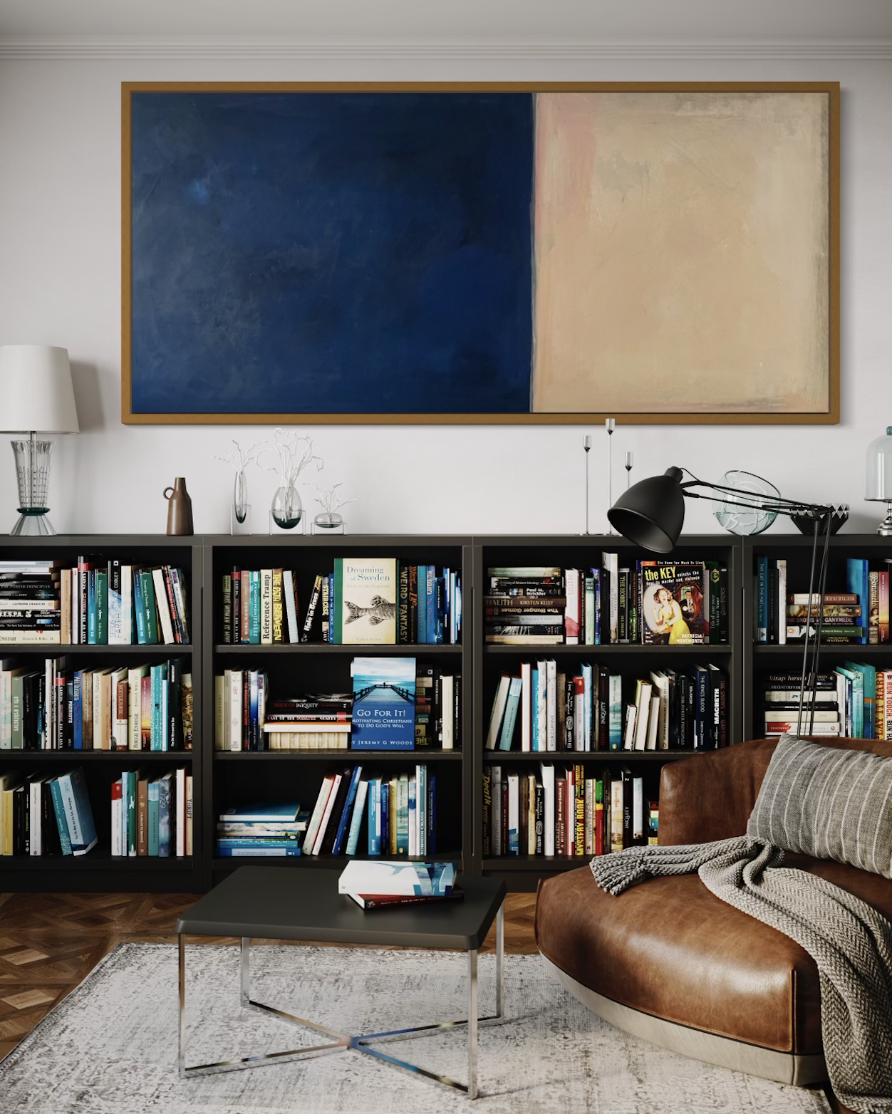 Living room with tall bookshelf filled with books, a brown leather sofa with a blanket, a small black coffee table, a large abstract painting with blue and beige sections, and a white wall.
