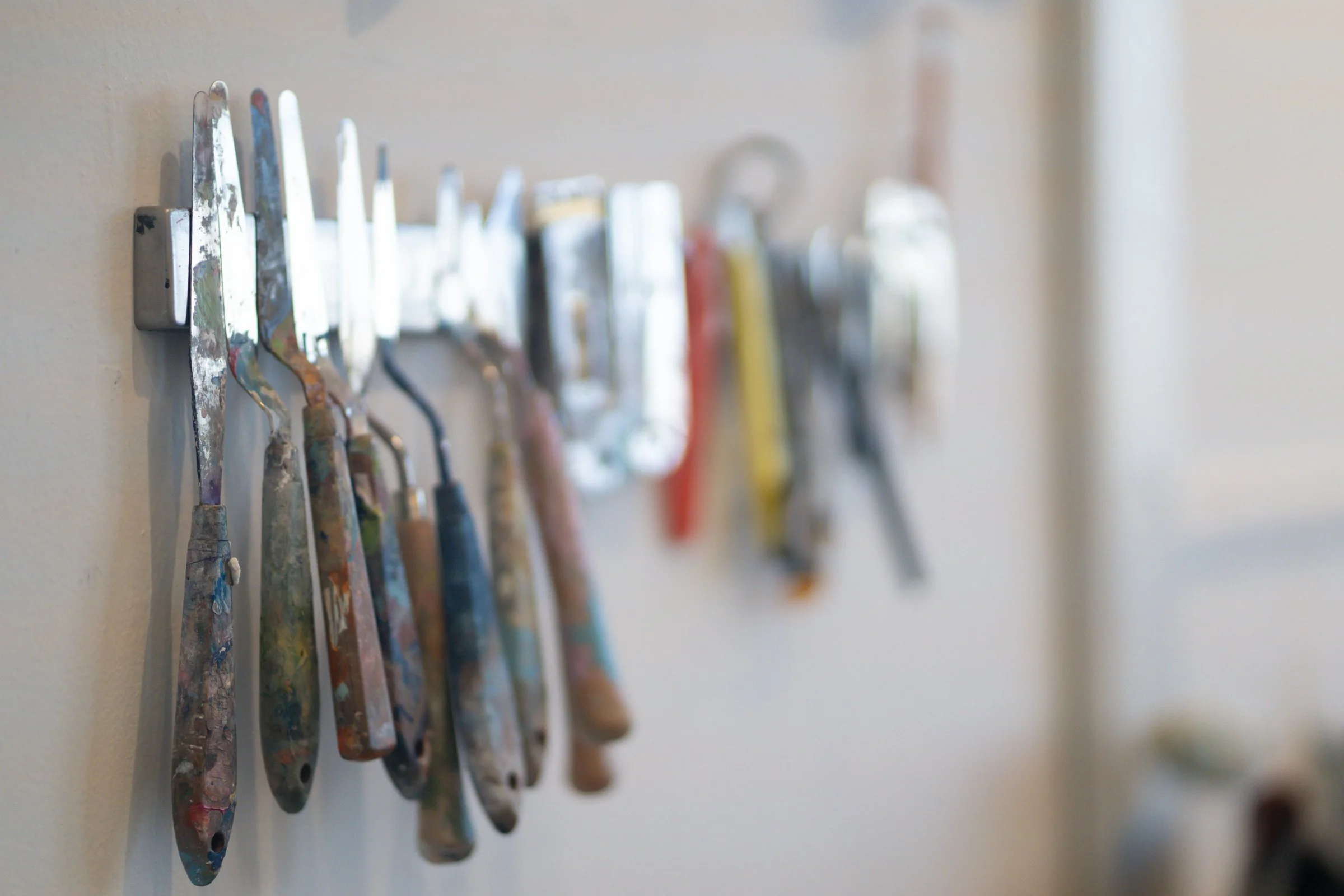 Paint palette knives hanging on a wall with some visible paint splatters.