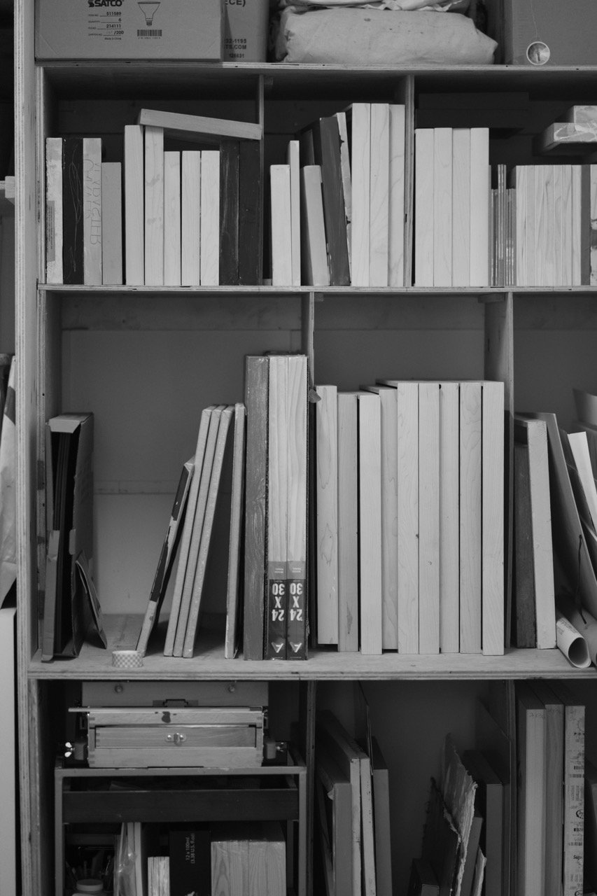 Black and white photo of a bookshelf filled with various books, magazines, and boxes, some leaning and stacked.