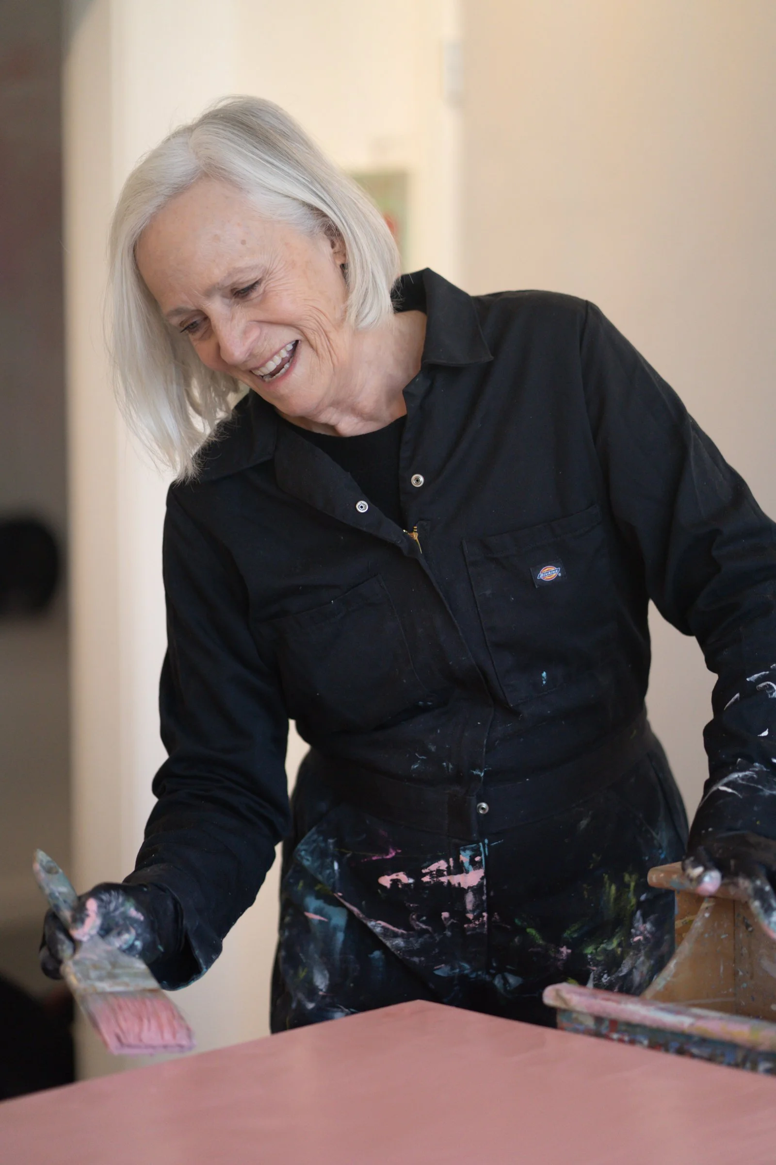 An elderly woman with white hair smiling while painting on a piece of pink paper, wearing a black work shirt and smock with paint stains.