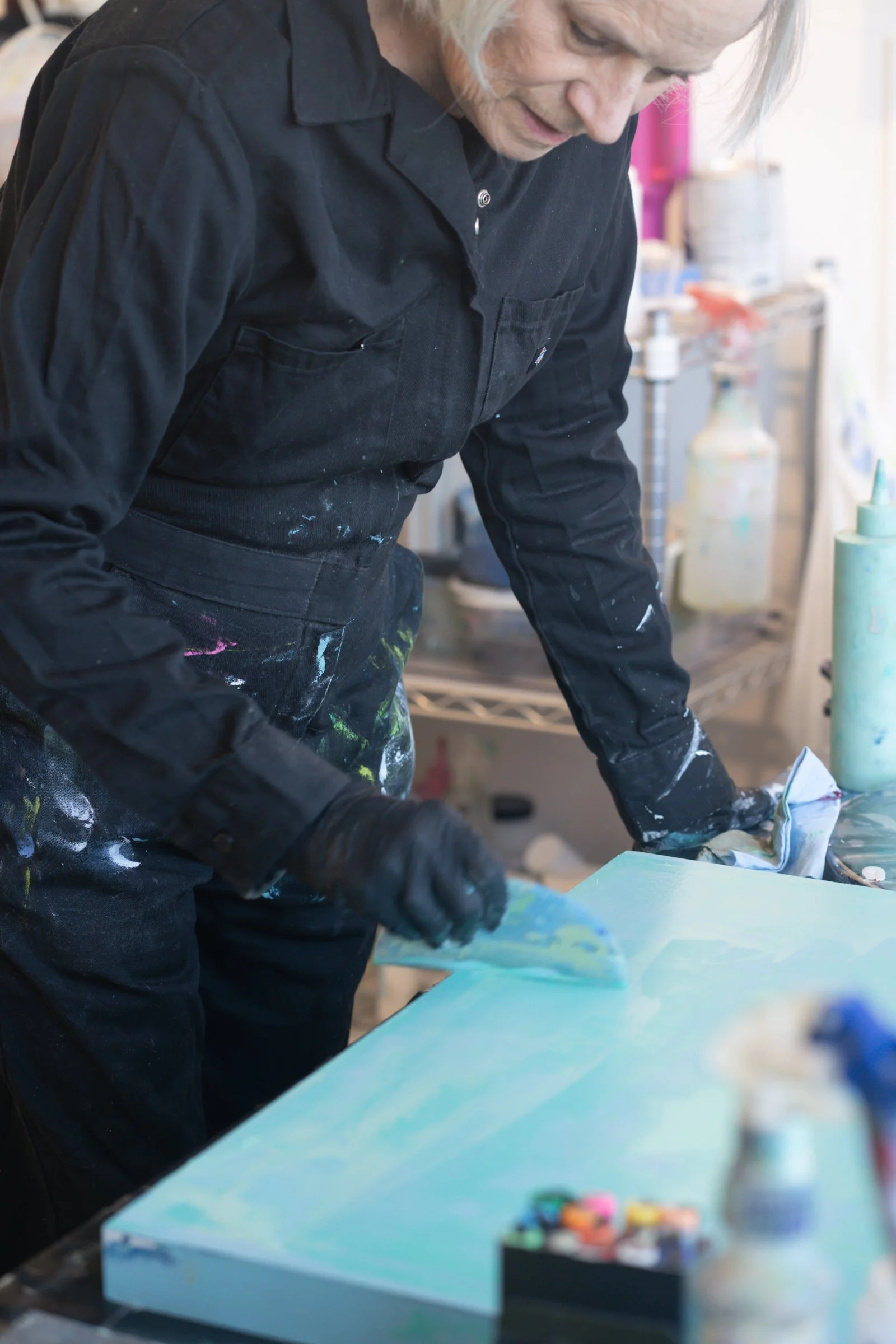 An elderly person wearing a black paint-splattered jumpsuit and gloves is applying paint to a canvas with a spatula. The workspace has art supplies and bottles in the background.