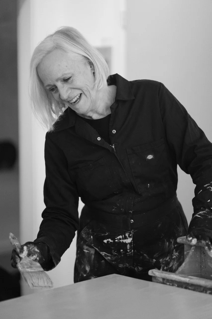 An elderly woman with blonde hair, smiling, wearing a black shirt and work apron, appears to be painting or working on a project at a table.