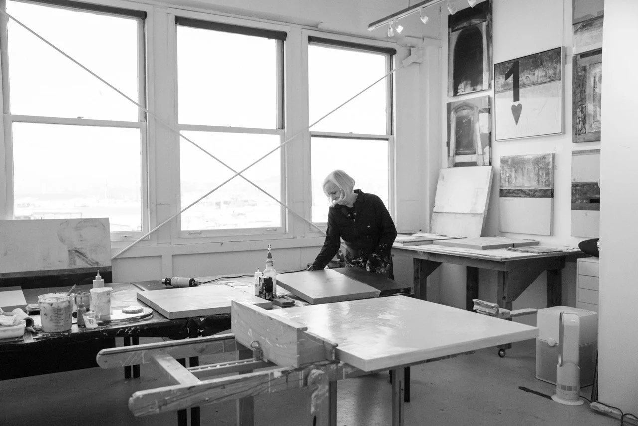 An artist working in a studio surrounded by paintings and art supplies, with large windows behind her and multiple artworks hanging on the wall.