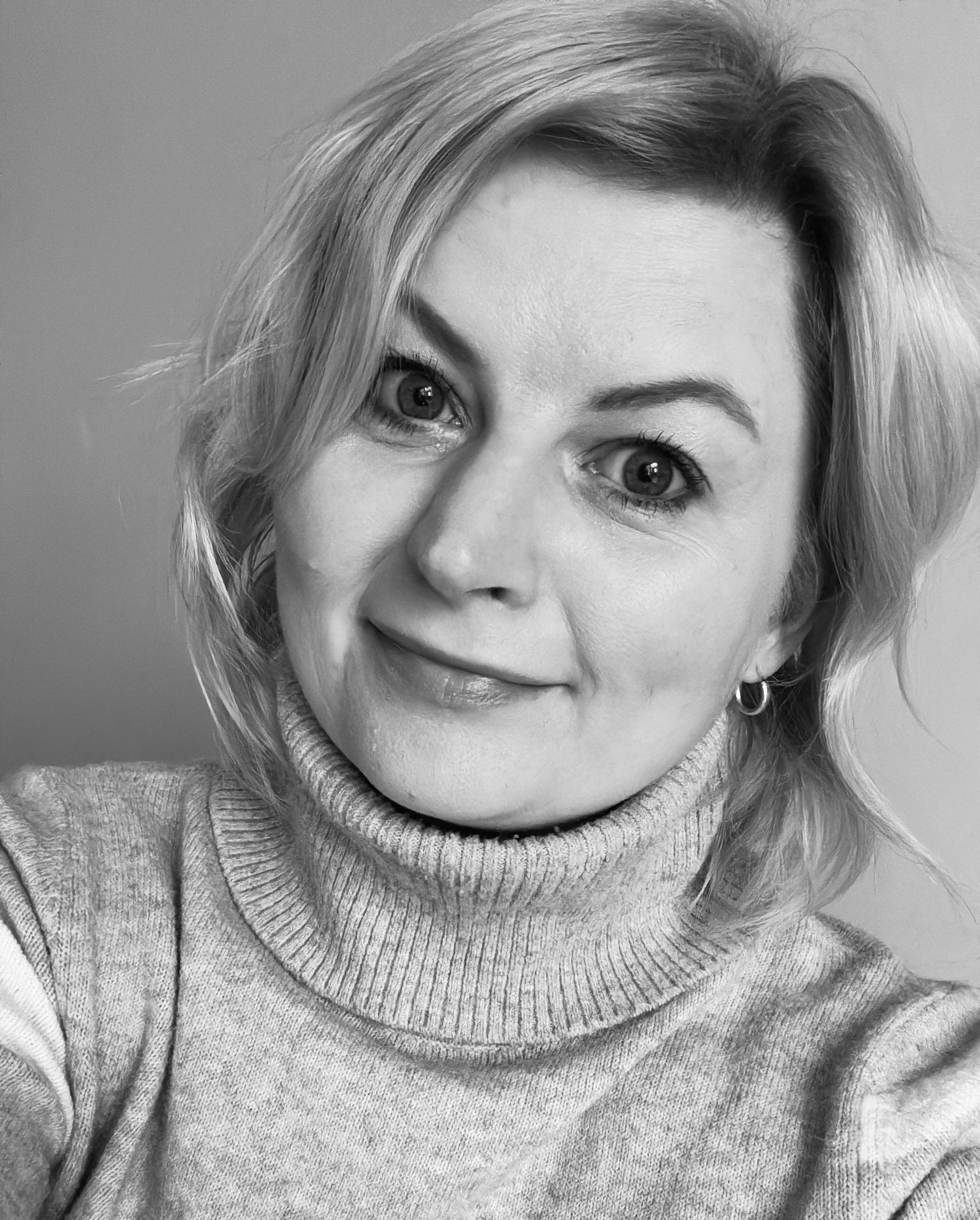 A woman with short, wavy blonde hair and earrings, smiling and wearing a turtleneck sweater.