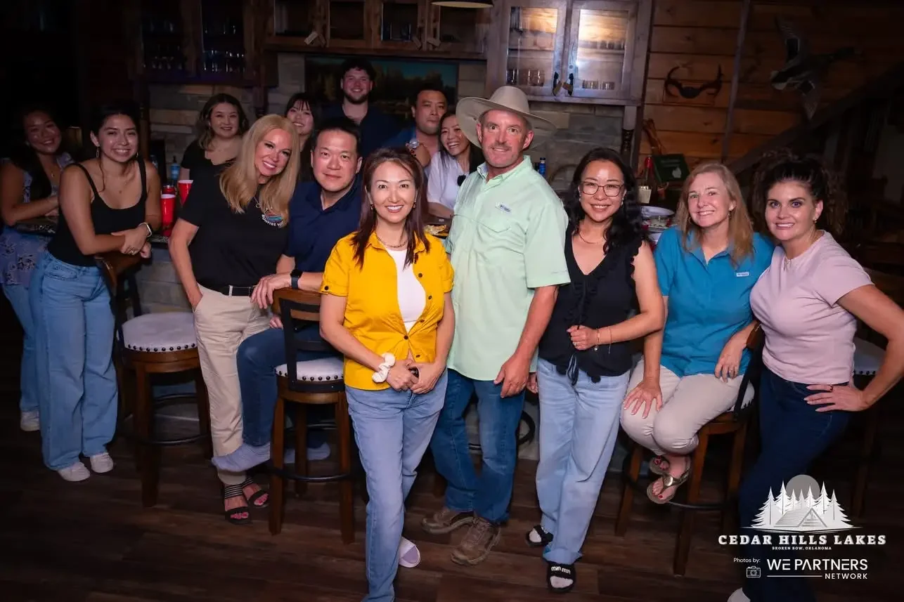 A group of people, mostly women, gathered in a rustic wood-paneled room, smiling for a photo around a bar counter. Some are seated while others are standing behind. There's a man in a light green shirt and wide-brimmed hat at the center. The background shows shelves with glassware and the logo for Cedar Hills Lakes in Oklahoma.