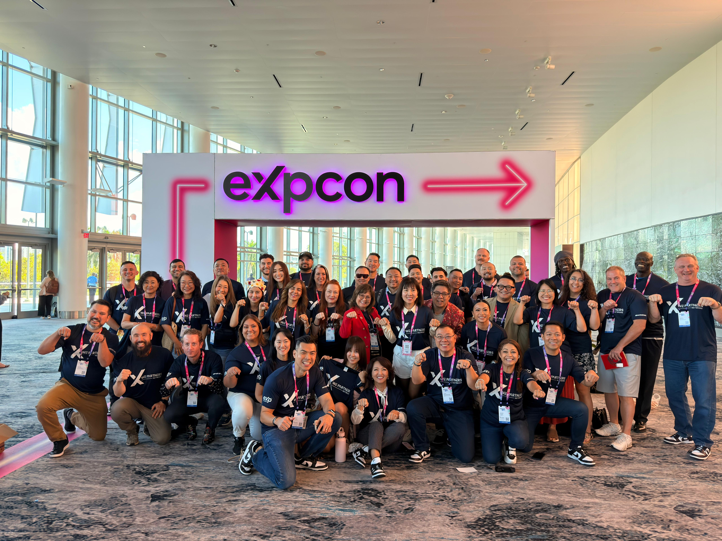 Large group of conference attendees posing together in front of a sign that reads 'expcon' with an arrow pointing to the right, inside a modern, glass-walled building with natural light.