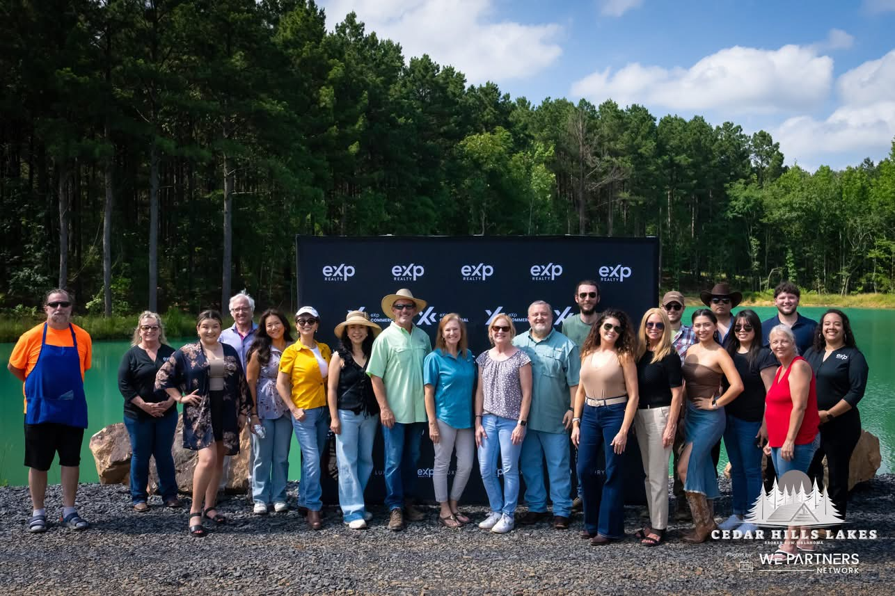 Group of people standing outdoors near a lake with trees in the background, posing for a photo in front of a black backdrop with the logo 'exp' and 'Cedar Hills Lakes' at the bottom right.