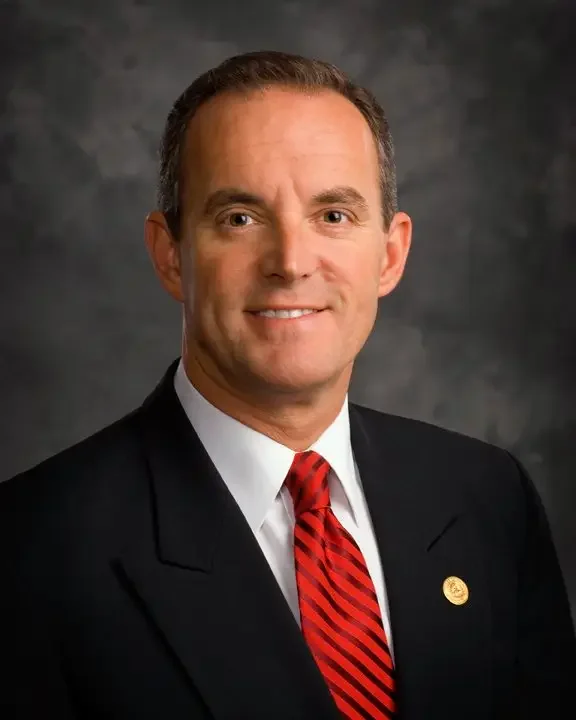A man wearing a black suit, white shirt, and a red striped tie, posing in front of a neutral gray background.
