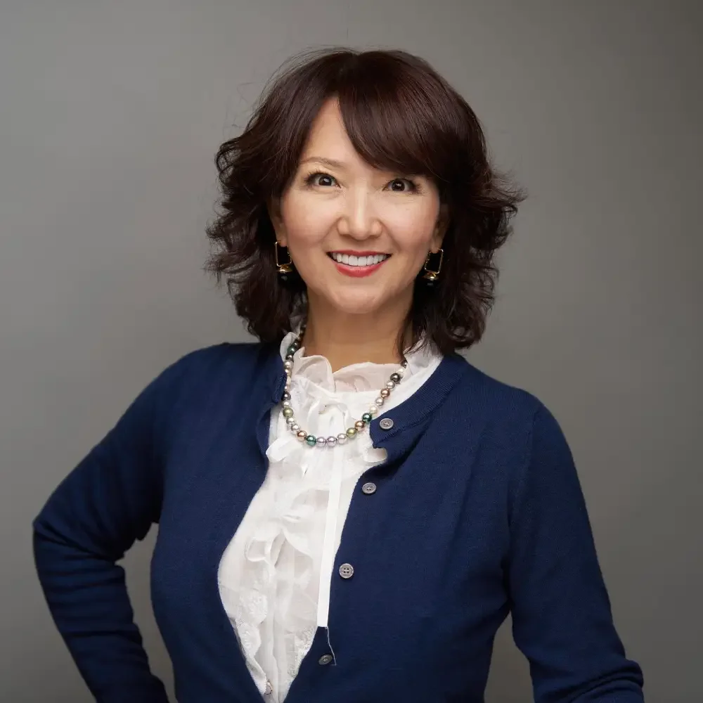 A woman with shoulder-length wavy brown hair, wearing a navy blue cardigan over a white blouse with ruffles, a multi-colored pearl necklace, and gold earrings, smiling against a neutral gray background.