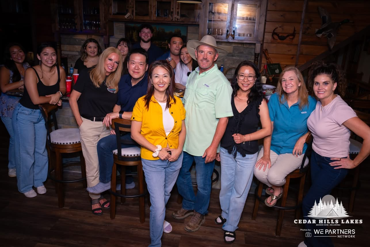 A group of people gathered together in a rustic indoor setting, some sitting on bar stools and others standing, smiling at the camera. There is a fireplace and wooden walls in the background. The image includes diverse individuals, some wearing casual clothing and one man with a wide-brim hat. Logo in the bottom right corner: Cedar Hills Lakes, Broken Bow, Oklahoma, and WE Partners Network.