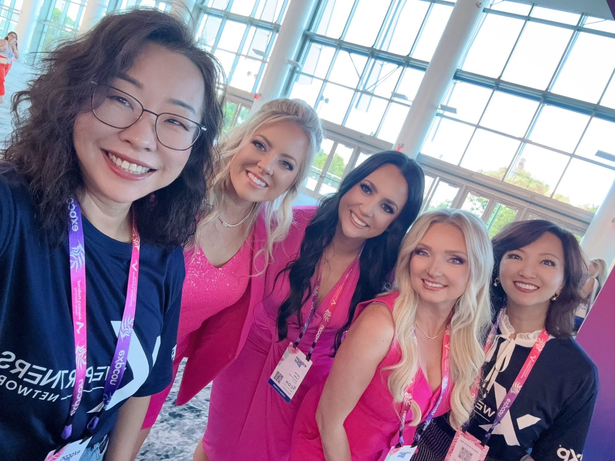 Group of women smiling at event, wearing pink and dark blue clothing, with large glass windows in background.