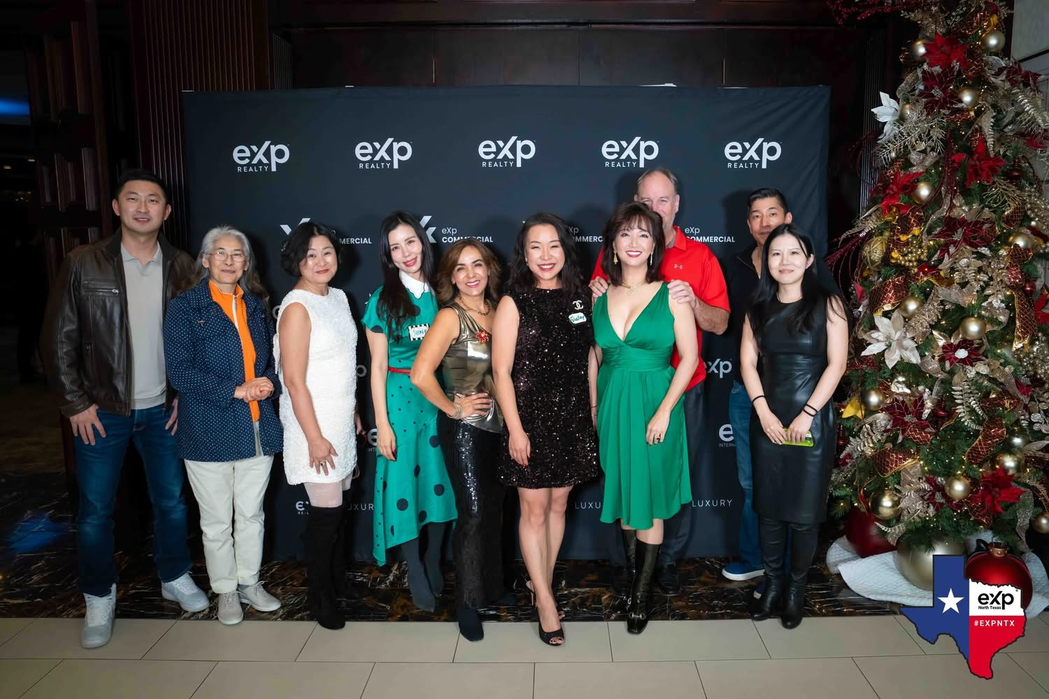 Group of people posing for a photo in front of a black backdrop with 'eXp Realty' logos, standing next to a decorated Christmas tree with red, gold, and white ornaments, during a holiday event in Texas.