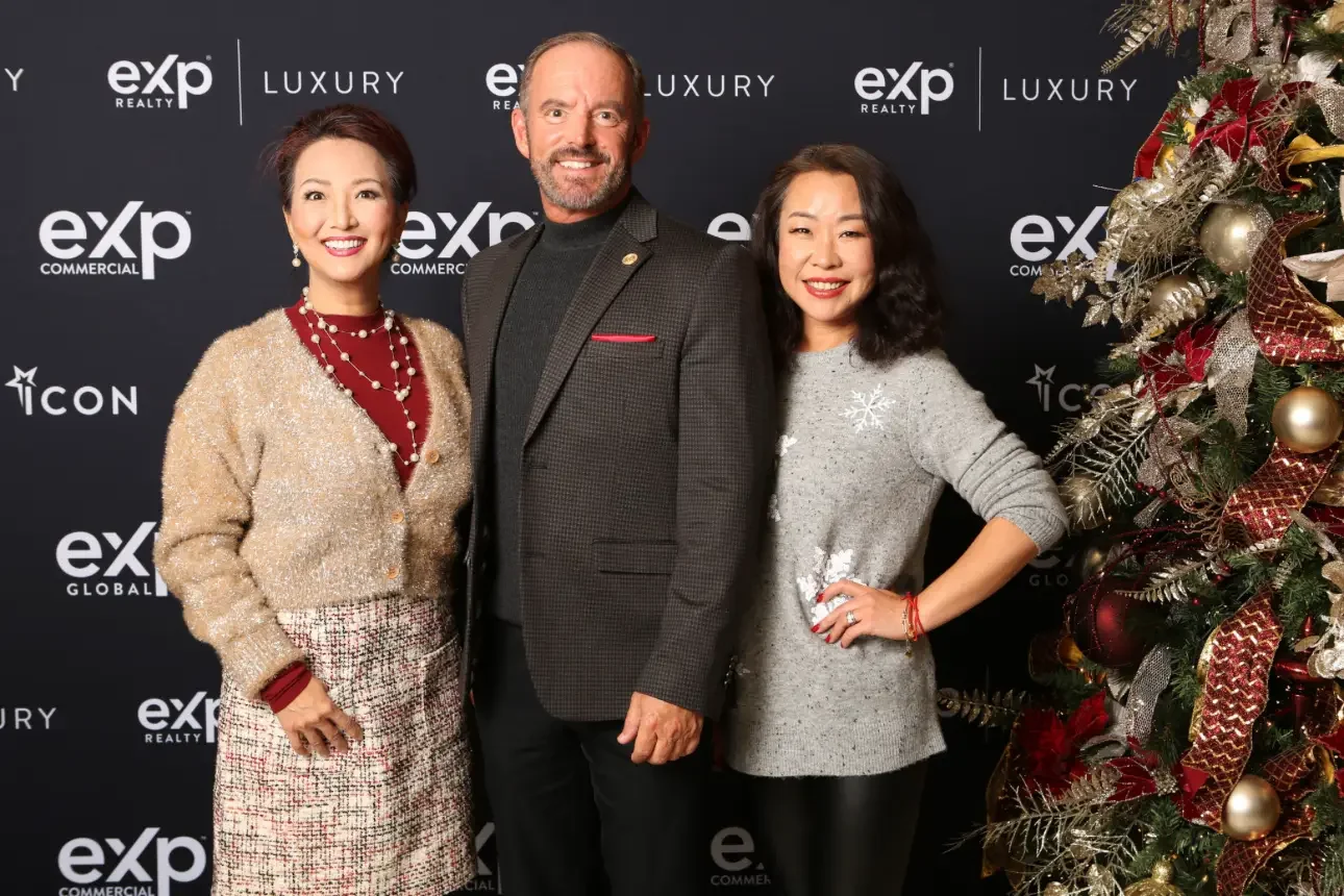 Three people standing together at a holiday event with a decorated Christmas tree on the right side and a black backdrop with logos for exp Realty, Luxury, and ICON on it.
