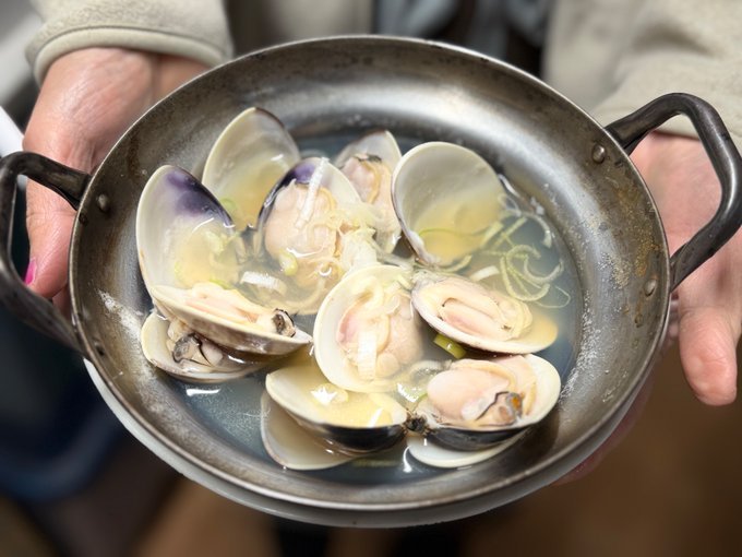 A person holding an iron dish containing clam broth with shells
