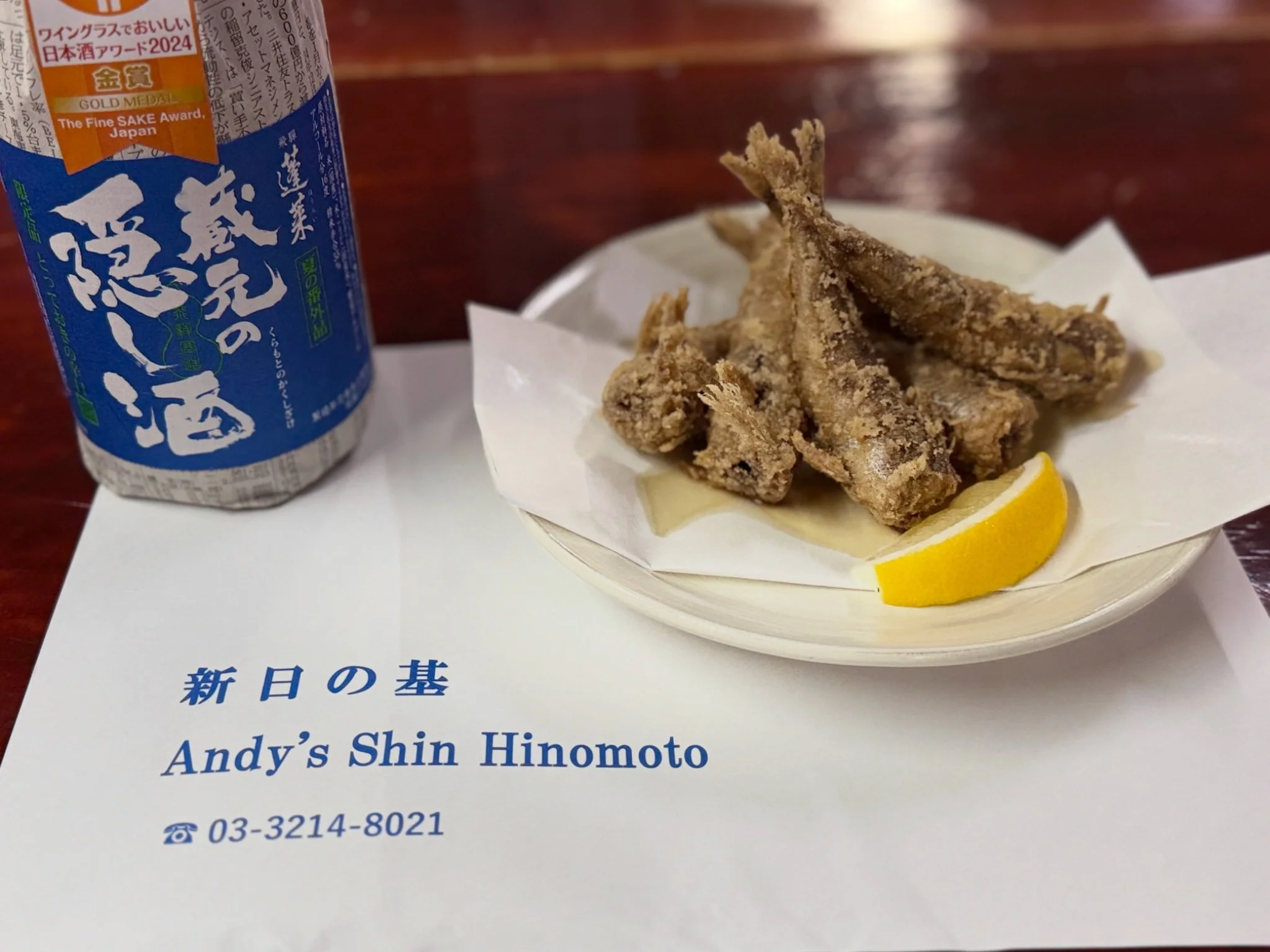 A photo of a table set with fried small fish, lemon slices, and a bottle of Japanese sake with a blue label.