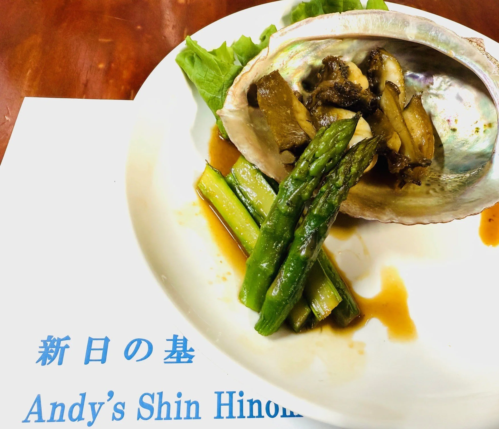 A dish served on a seashell, featuring asparagus and zucchini vegetables with sauce. The restaurant name is written in blue text in Japanese and English at the bottom left of the plate.