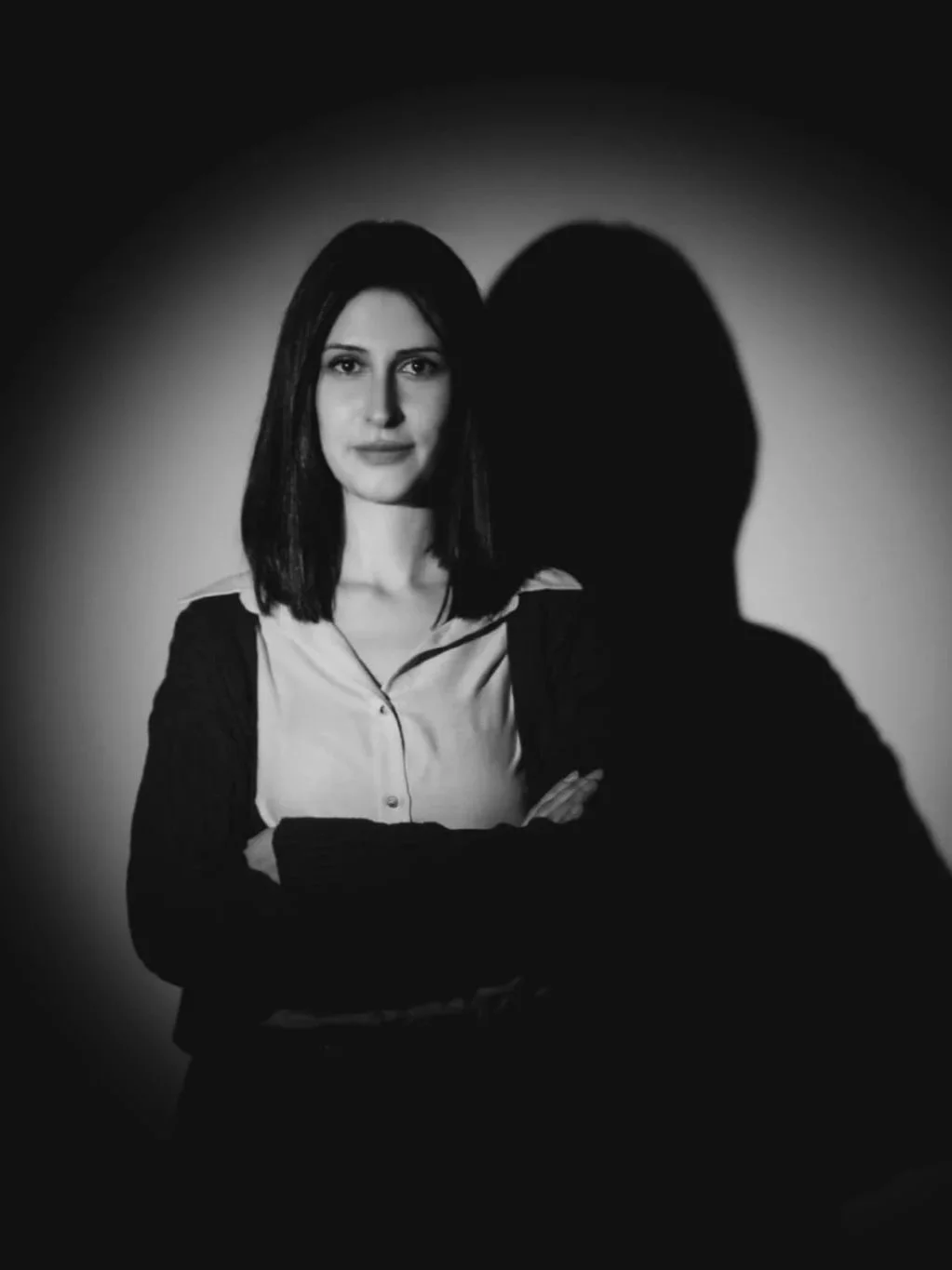 Black and white portrait of a young woman with dark hair, standing with arms crossed and looking at the camera. She is wearing a light shirt and a darker jacket, with a shadow cast on the wall behind her.