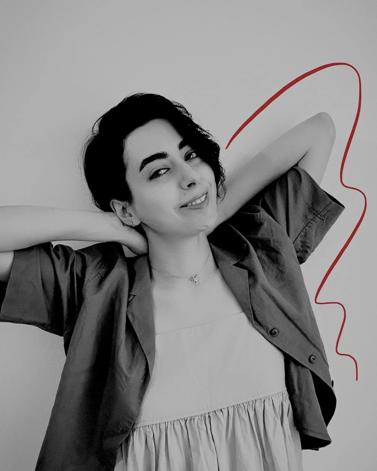 A woman with dark hair smiling, with her arms behind her head, wearing a light-colored dress and a dark open shirt, standing against a plain wall with red line art of a face outline in the background.