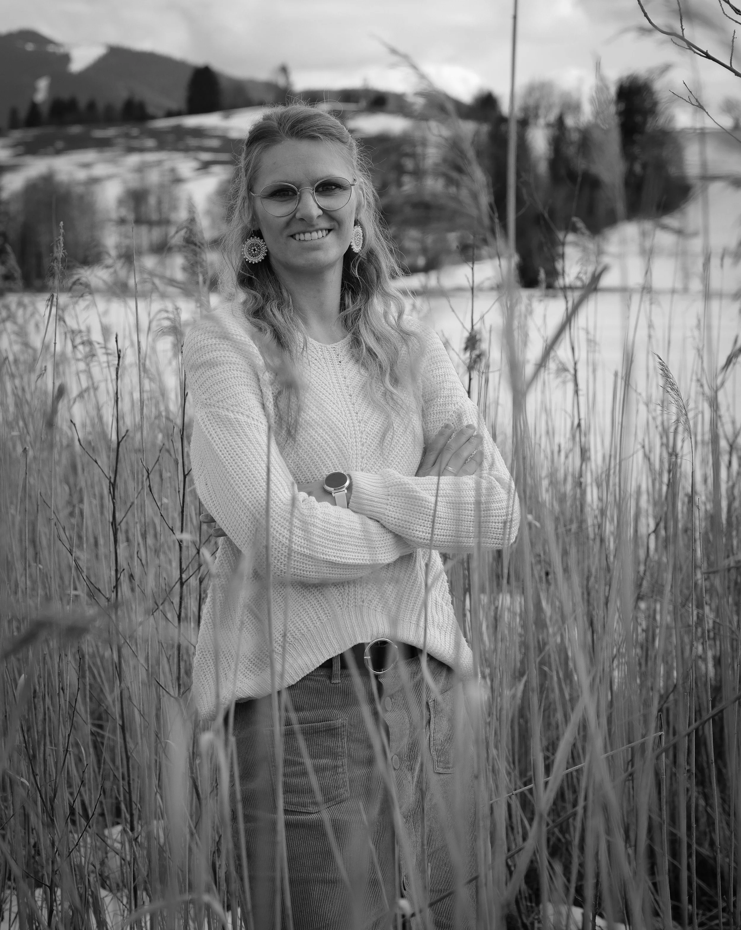 Schwarz-Weiß-Foto einer lächelnden Frau mit Brille in einem Kleid und Pullover, die in einer Wiese mit hohen Gräsern steht, im Hintergrund Berge und Bäume.