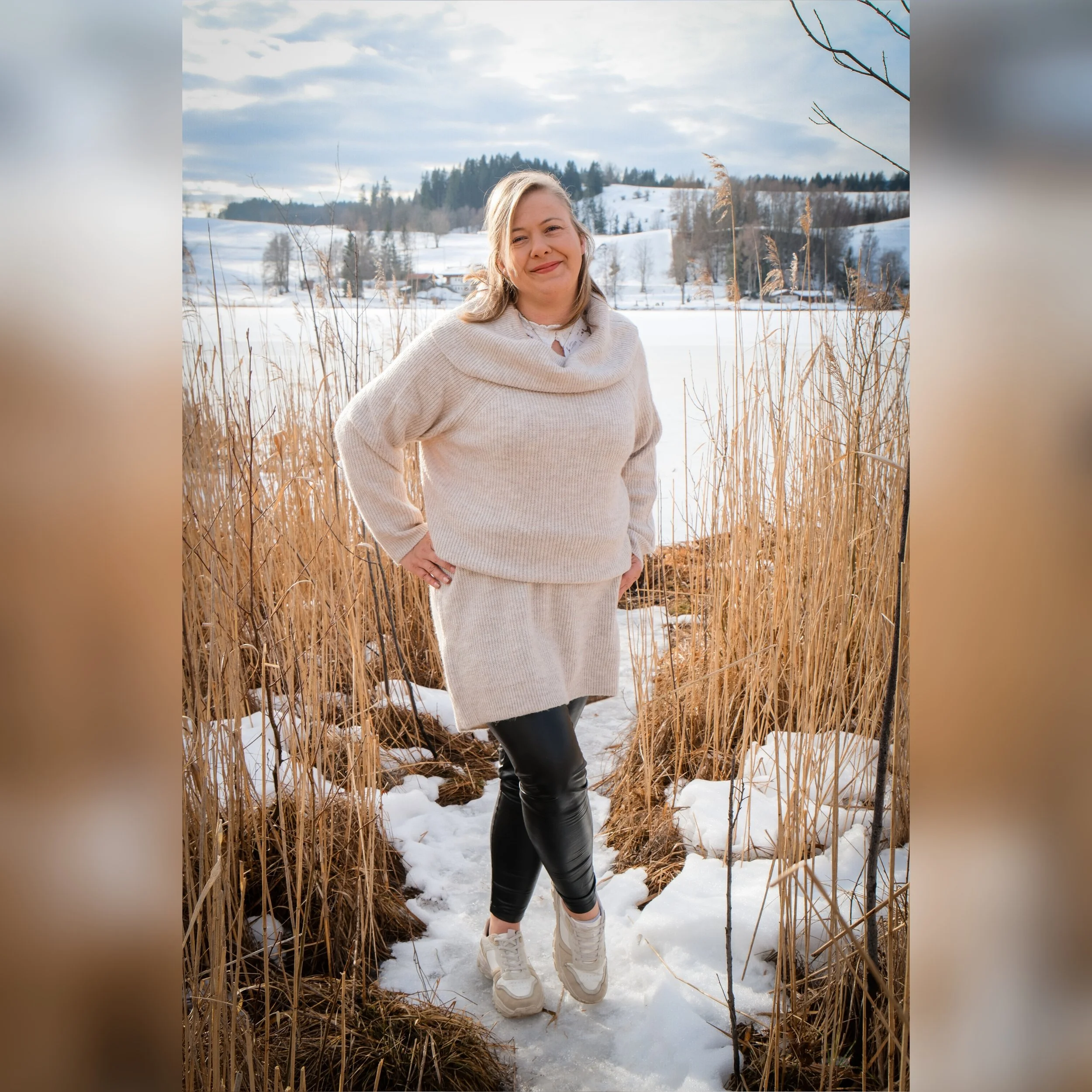 Eine Frau steht in einer verschneiten Landschaft mit hohen, braunen Gräsern um sie herum und einem gefrorenen See im Hintergrund. Es ist bewölkt, und im Hintergrund sind verschneite Hügel und Bäume sichtbar.