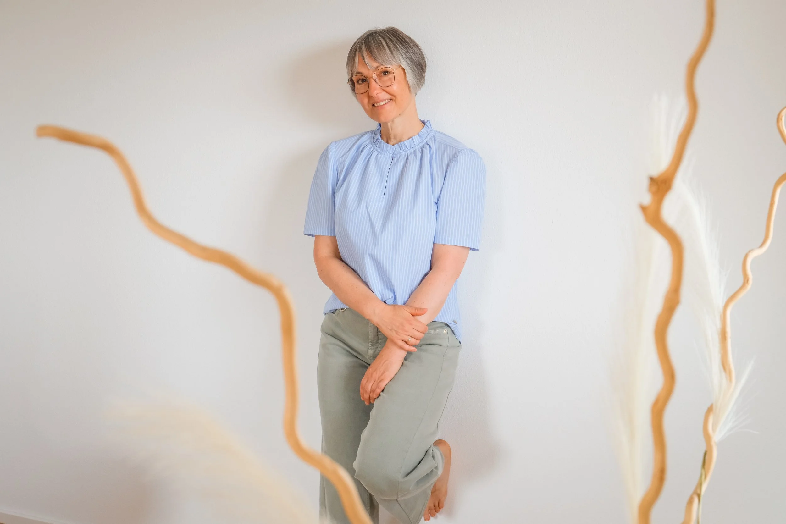 Eine Frau steht in einer verschneiten Landschaft mit hohen, braunen Gräsern um sie herum und einem gefrorenen See im Hintergrund. Es ist bewölkt, und im Hintergrund sind verschneite Hügel und Bäume sichtbar.