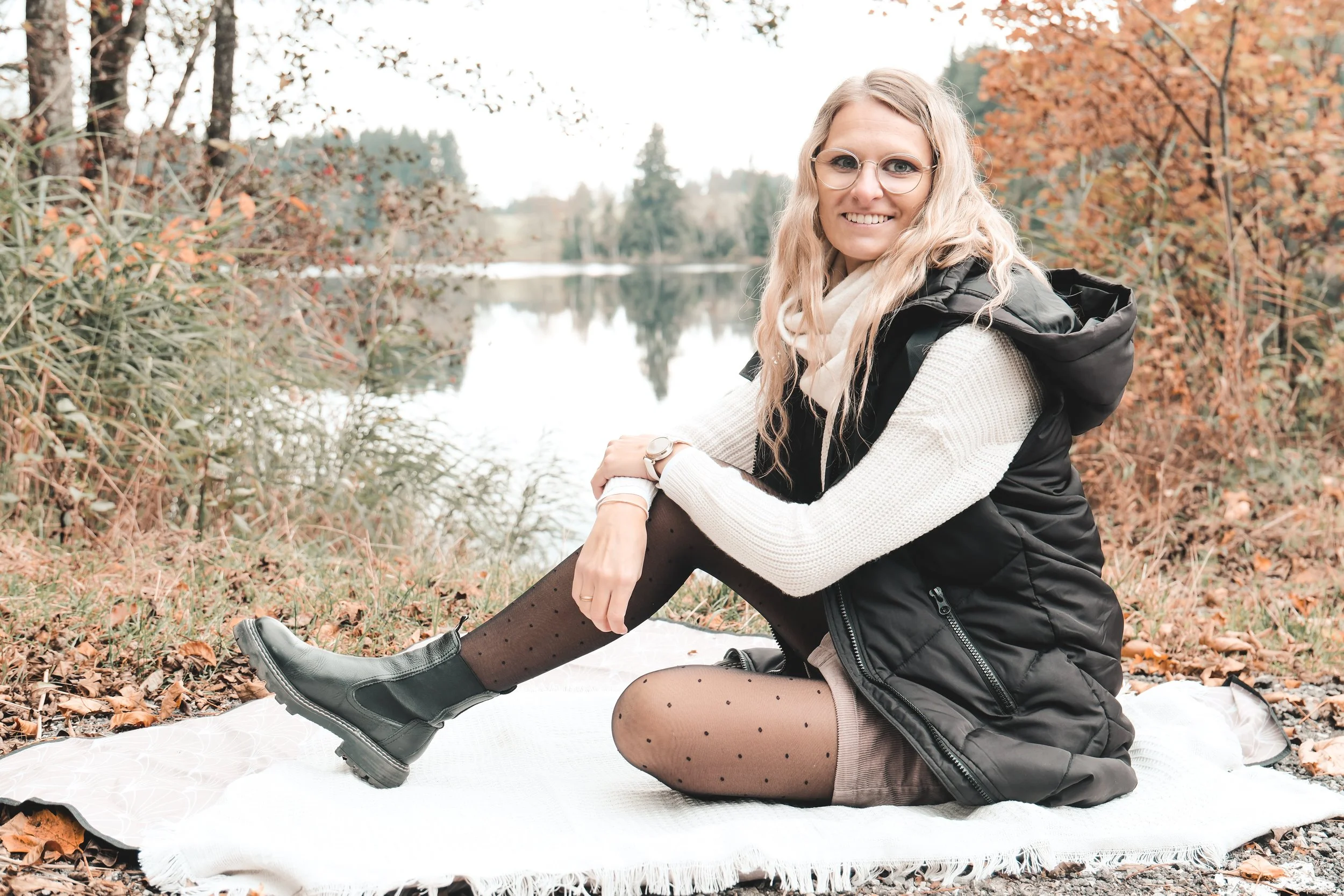 Eine lachende Frau mit Brille sitzt auf einer Decke am Fluss im Herbst, trägt einen schwarzen Weste, eine weiße Strickjacke, bunte Röhrenhose, Strumpfhosen mit Punkten und schwarze Stiefel.