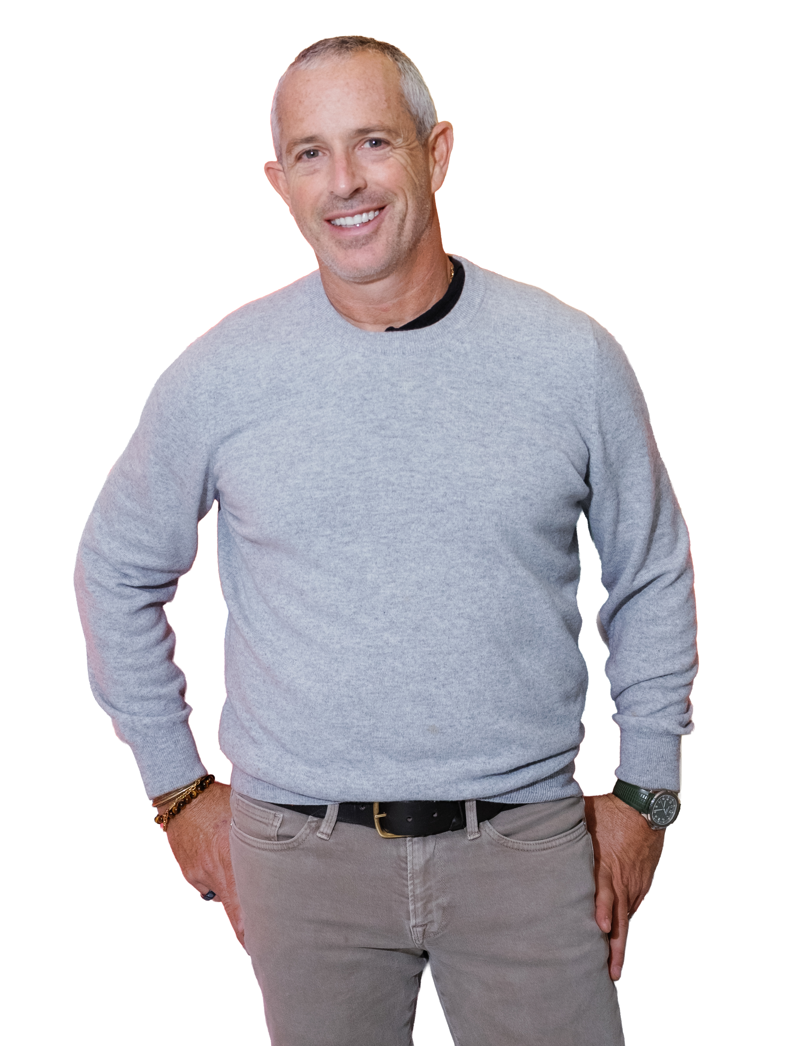 A middle-aged man with short gray hair, smiling, wearing a light gray sweater, beige pants, and a black belt, standing with one hand in his pocket.
