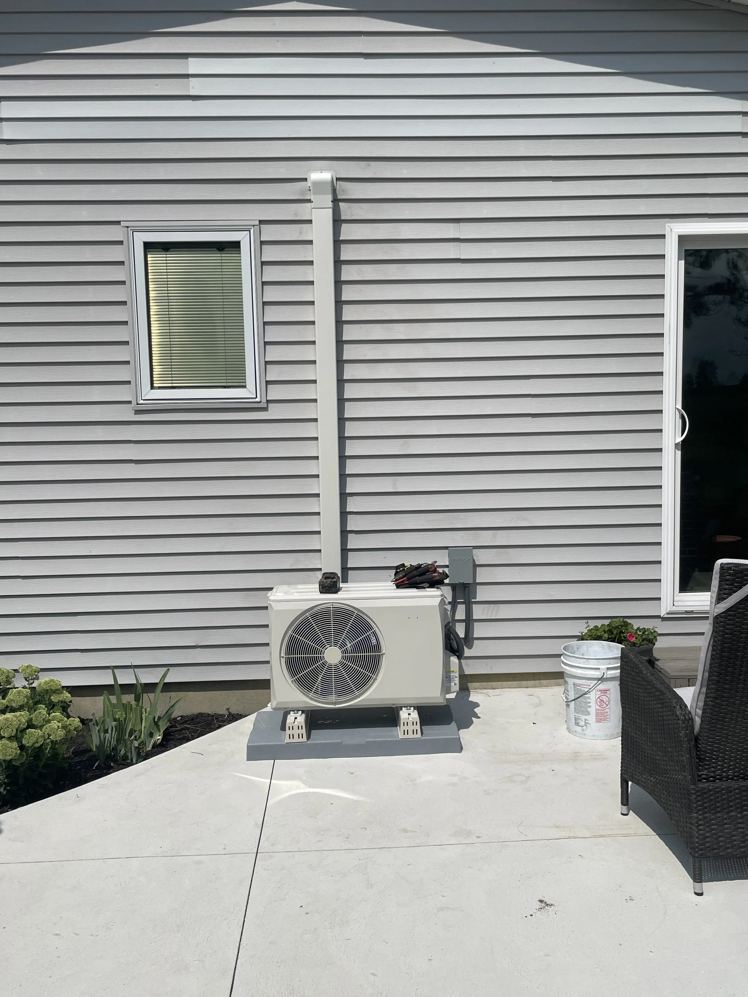 Outside view of a house with gray siding, a small window with blinds, a sliding glass door, an air conditioning unit on a concrete patio, a white bucket, a potted plant, and a black outdoor chair.