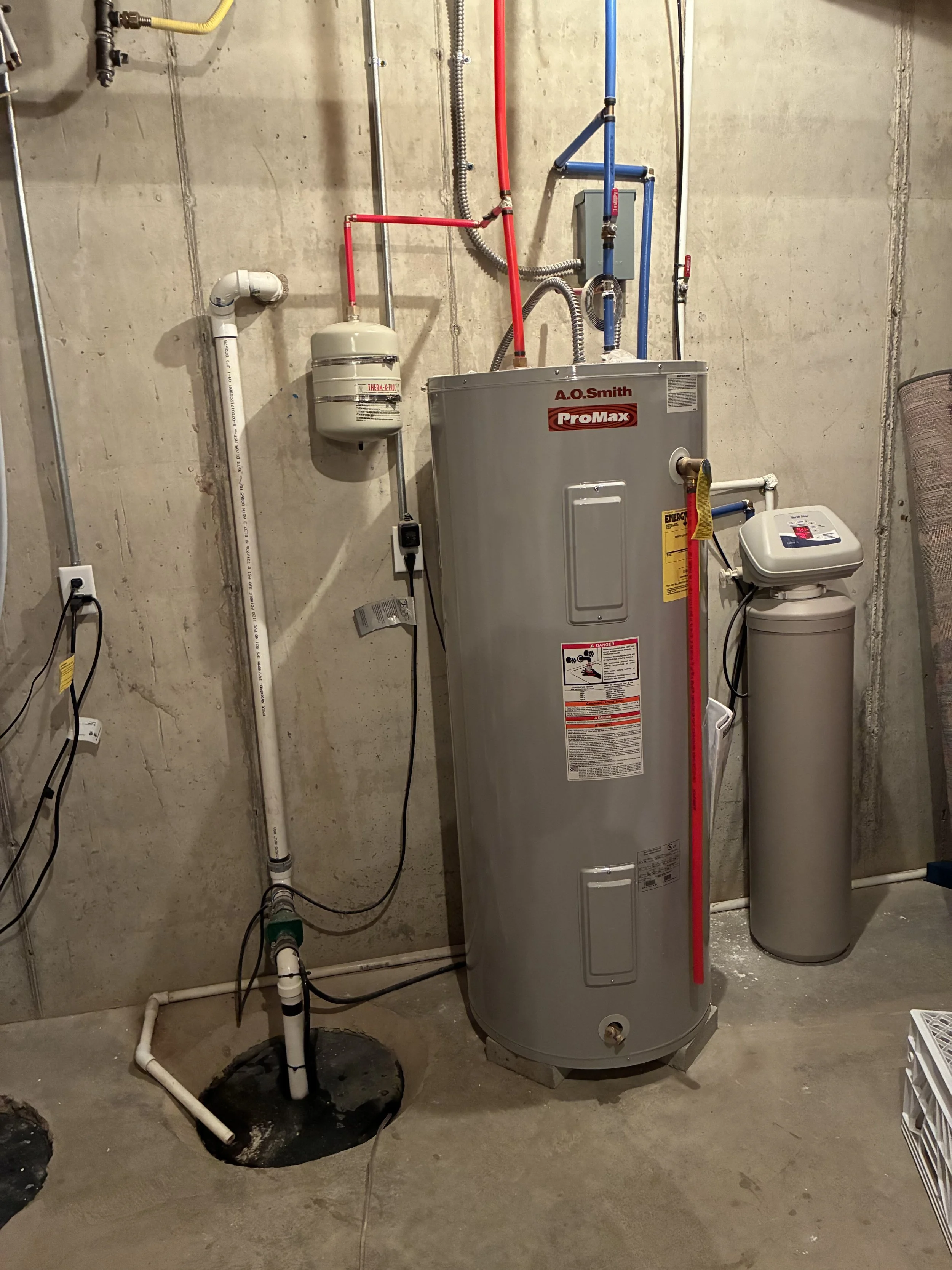 A utility room with a water heater, water softener, and plumbing pipes against a concrete wall.