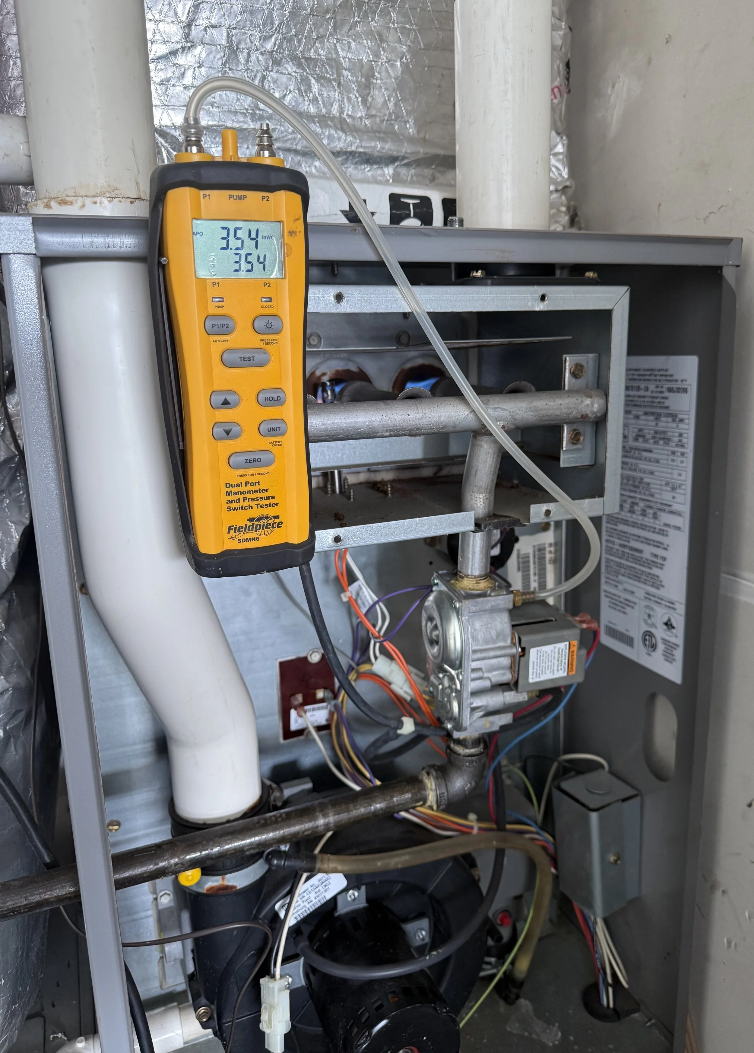 A yellow Dual Port Manometer and Pressure Switch Tester connected to an HVAC system, with the display showing readings of 3.54 and 3.54, inside an air handler or furnace with visible wiring, pipes, and metal components.