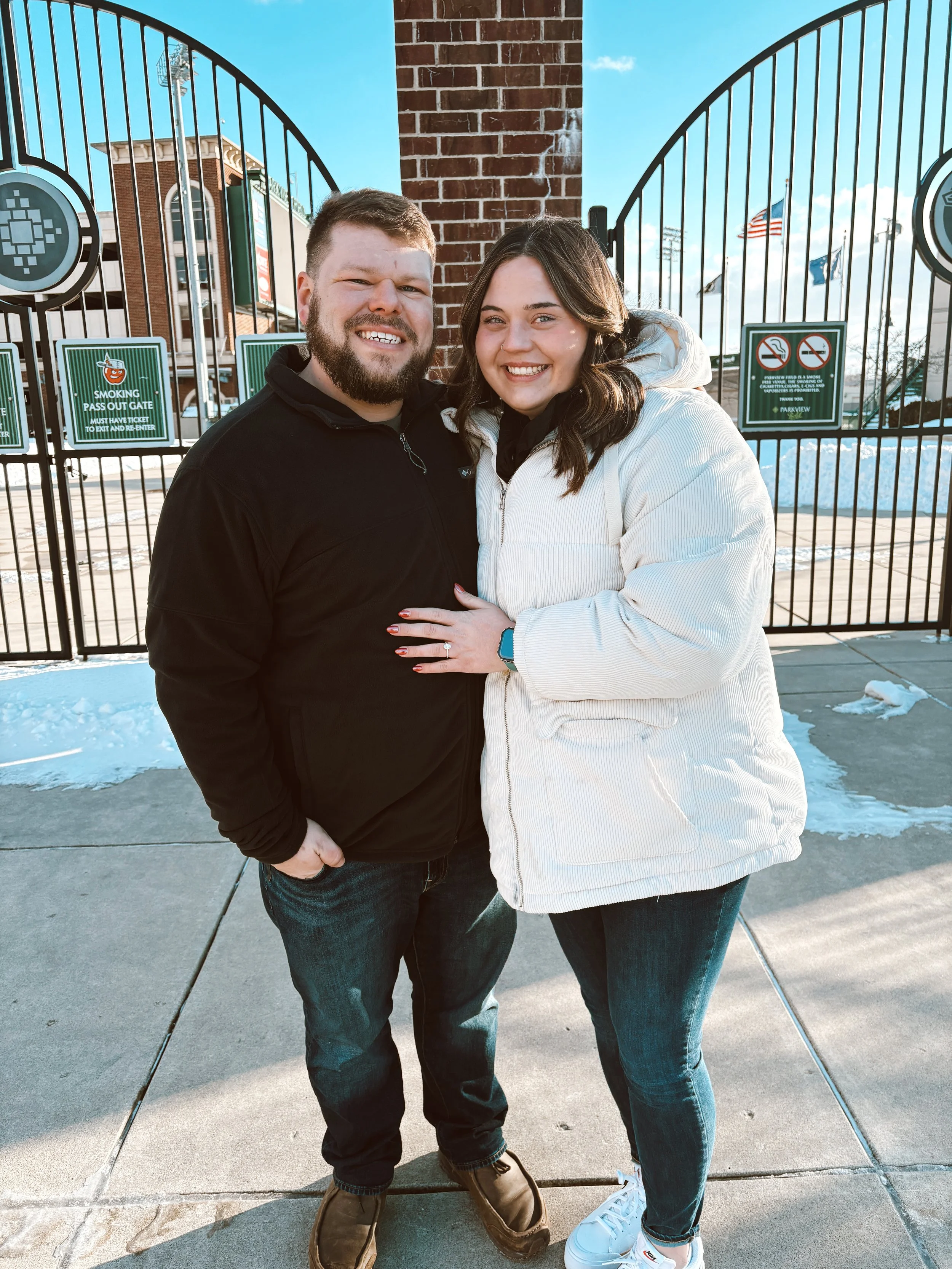 A smiling man and woman standing close together outdoors on a sidewalk, with a brick archway and gate behind them. Snow on the ground, some flags in the background.