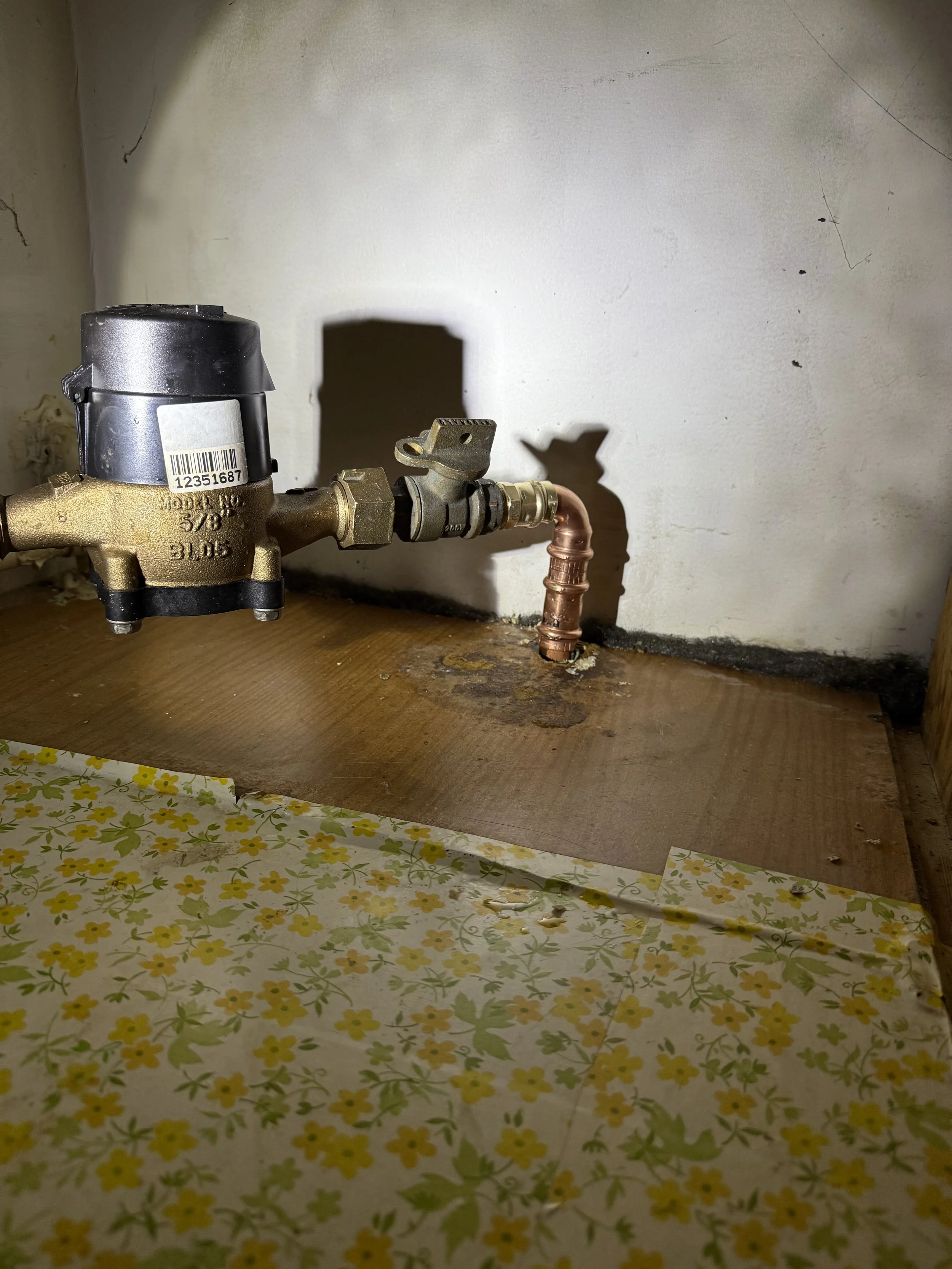 Plumbing pipe with a shut-off valve and copper pipe connection, on a wooden surface with yellow floral patterned linoleum.