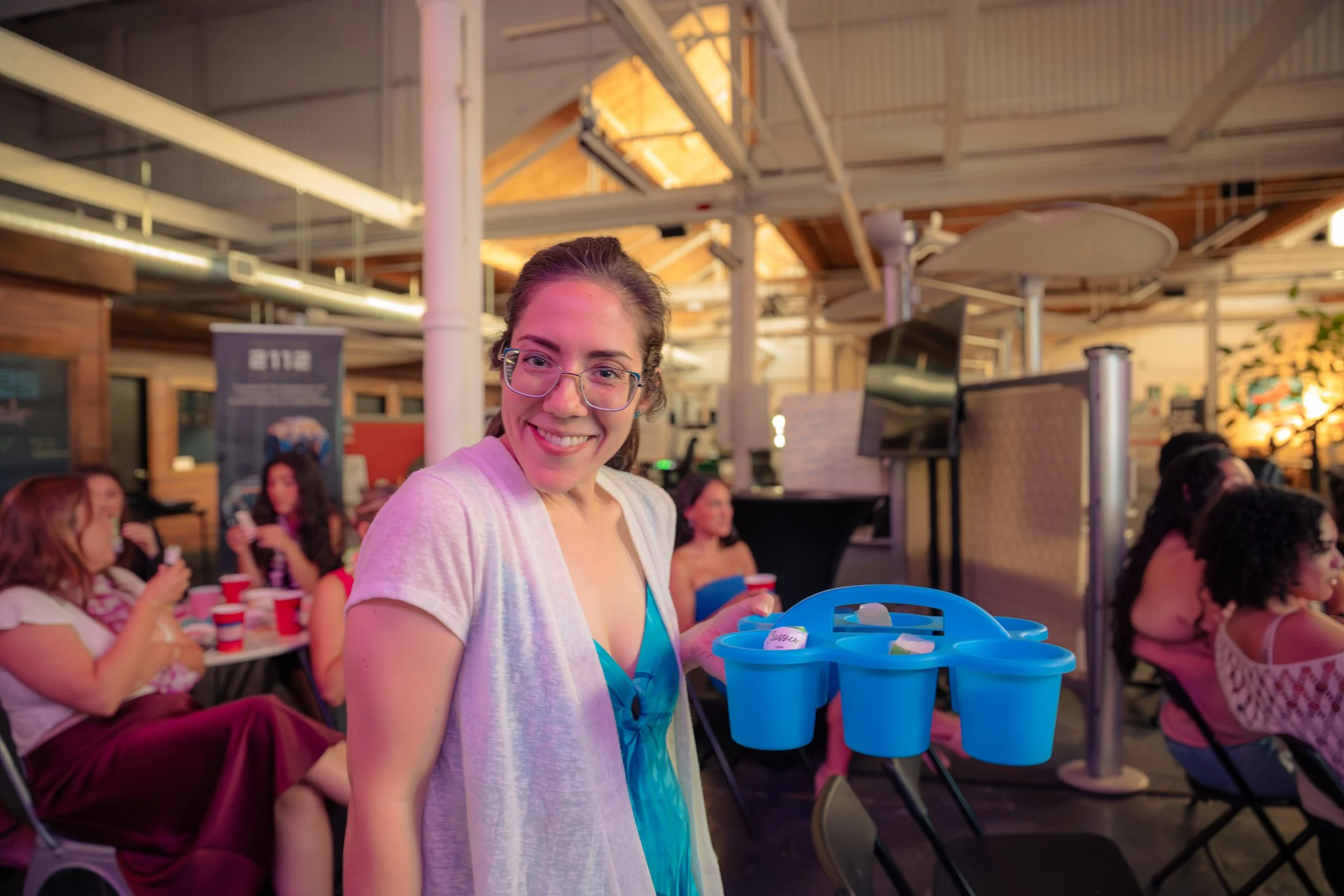 A smiling woman in glasses holding a blue tray with small containers inside at a social gathering in an indoor venue with other women seated at tables in the background.