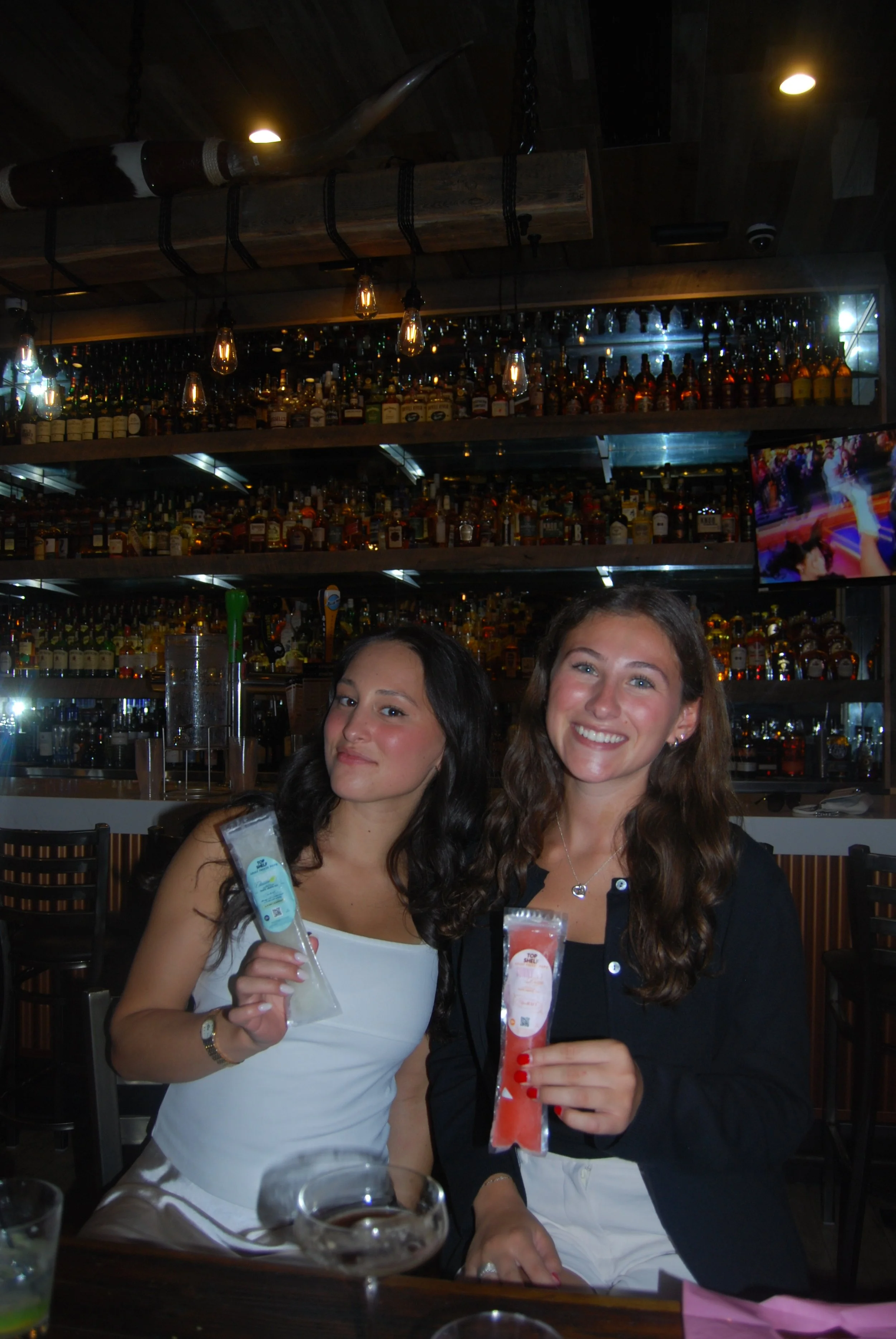 Two women sitting at a bar holding ice packs, one with a white top and the other with a black jacket, smiling for the camera, with a fully stocked bar in the background.
