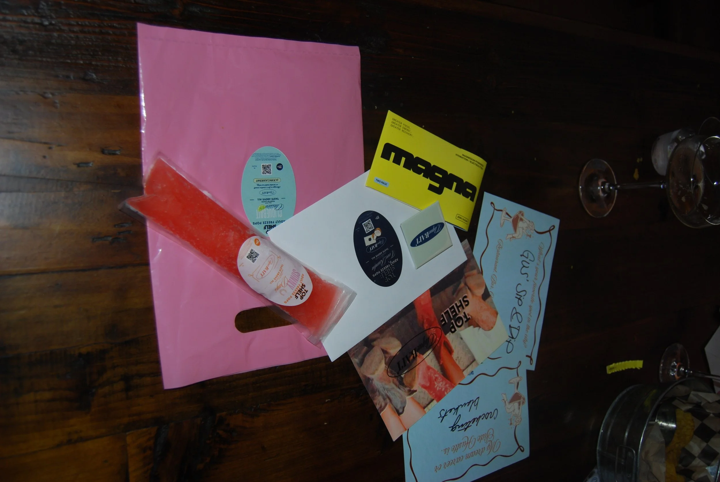 Various promotional items and signs on a wooden table, including a pink paper bag, a small packaged snack, a yellow sticker, and printed signs with text.
