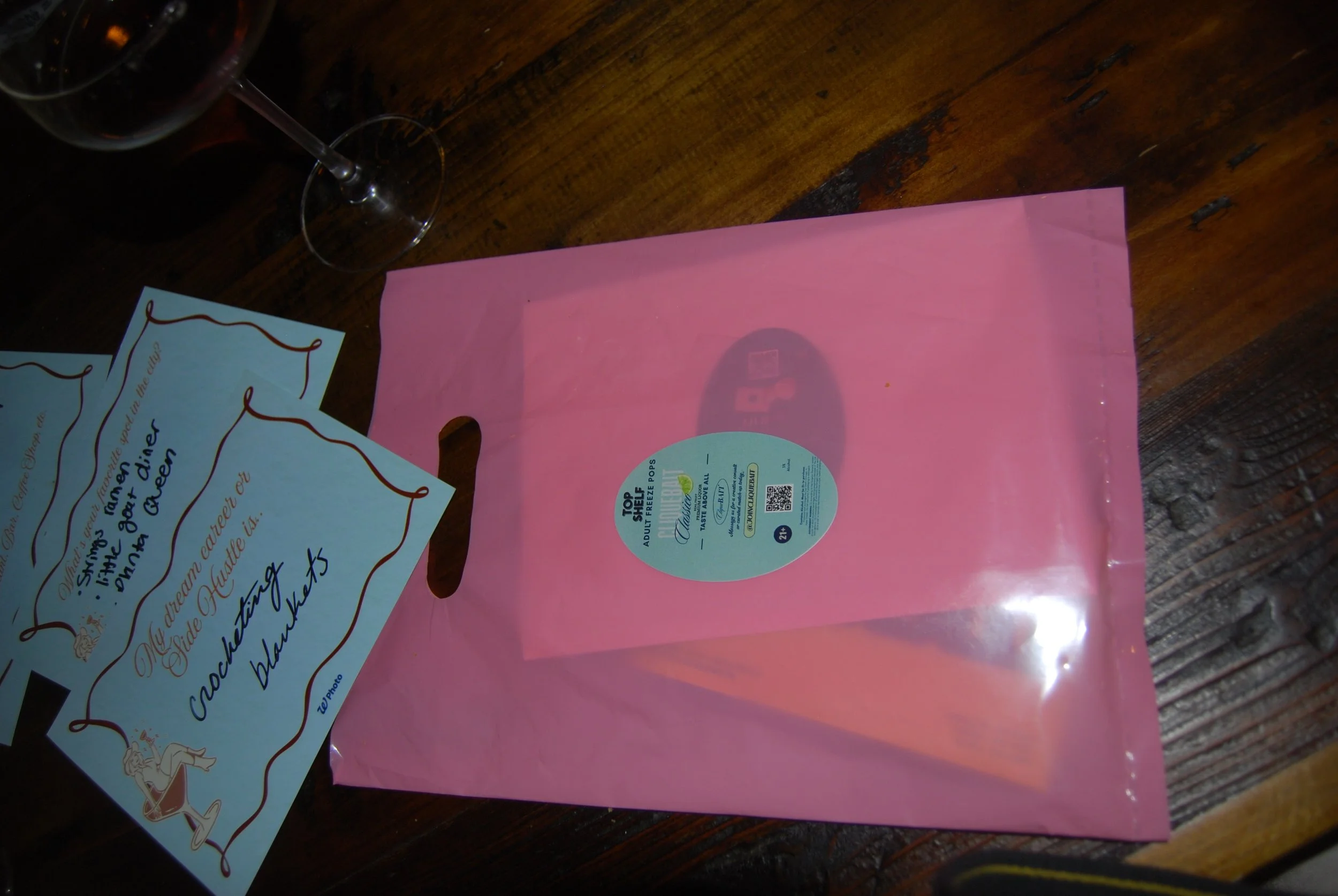 Pink plastic bag with a round label, partially transparent, placed on a wooden table. To the left, there are some cards with handwritten text and a glass of red wine.