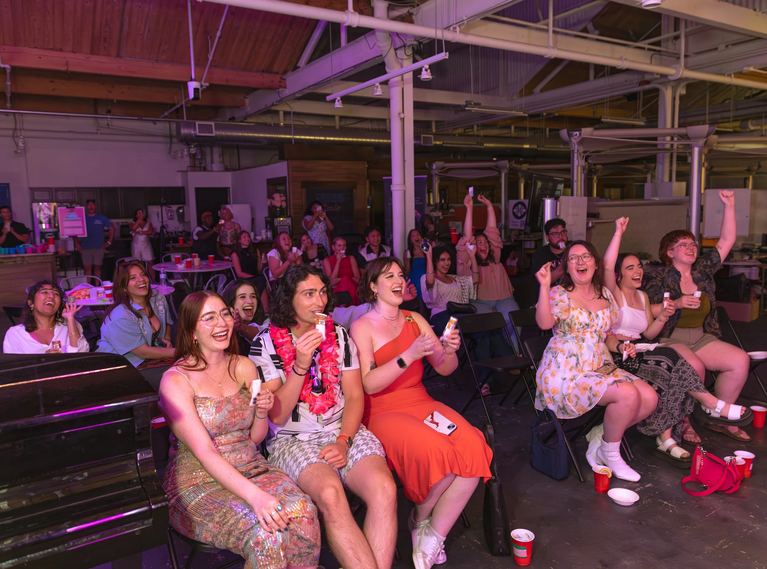 A group of people at an indoor event, sitting and enjoying a performance or show, with many raising their hands and smiling joyfully.