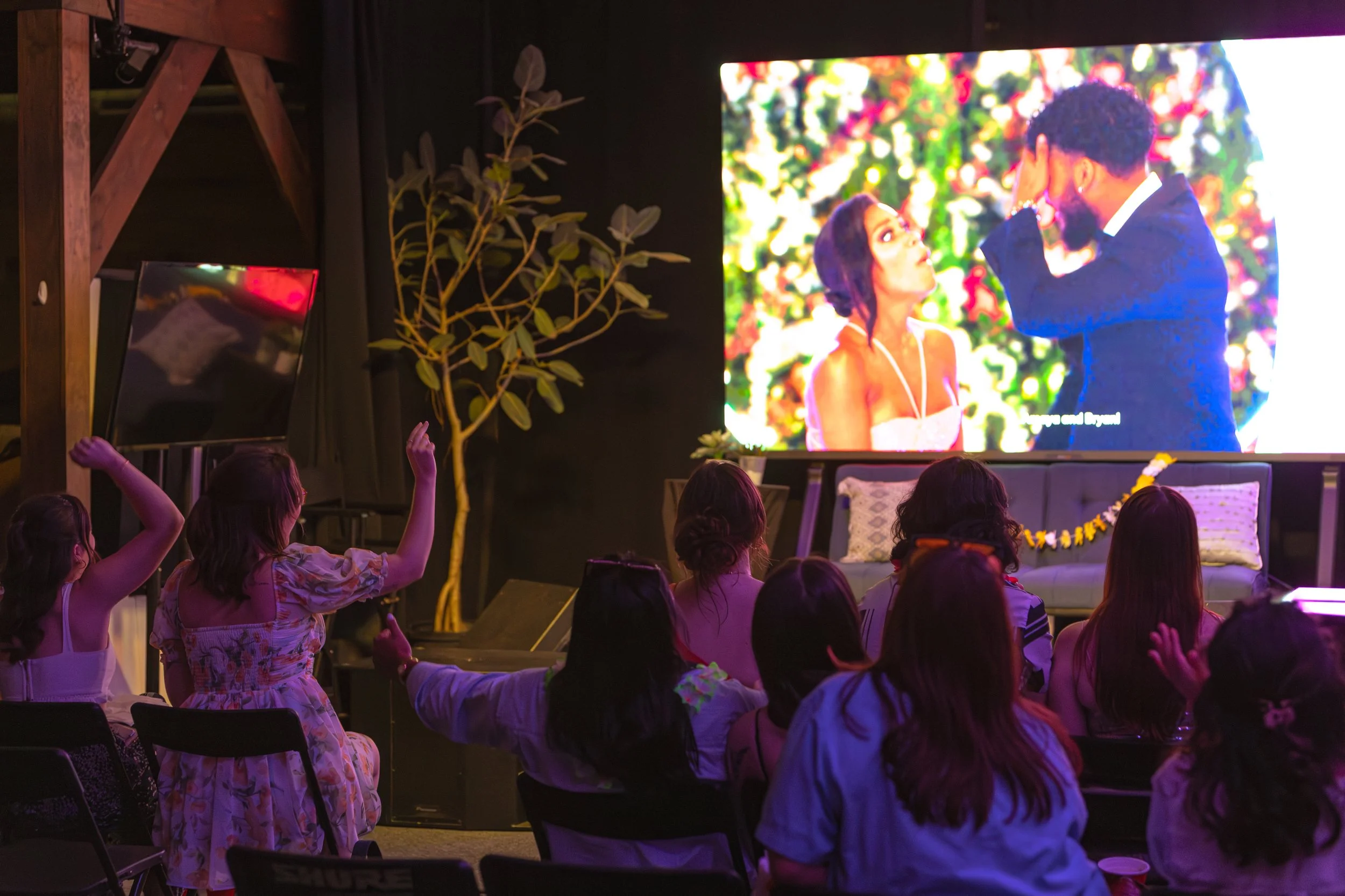 Women sitting in chairs watching a movie or show on a large screen in a dark room. The screen displays a scene with a man and a woman outdoors surrounded by colorful flowers.