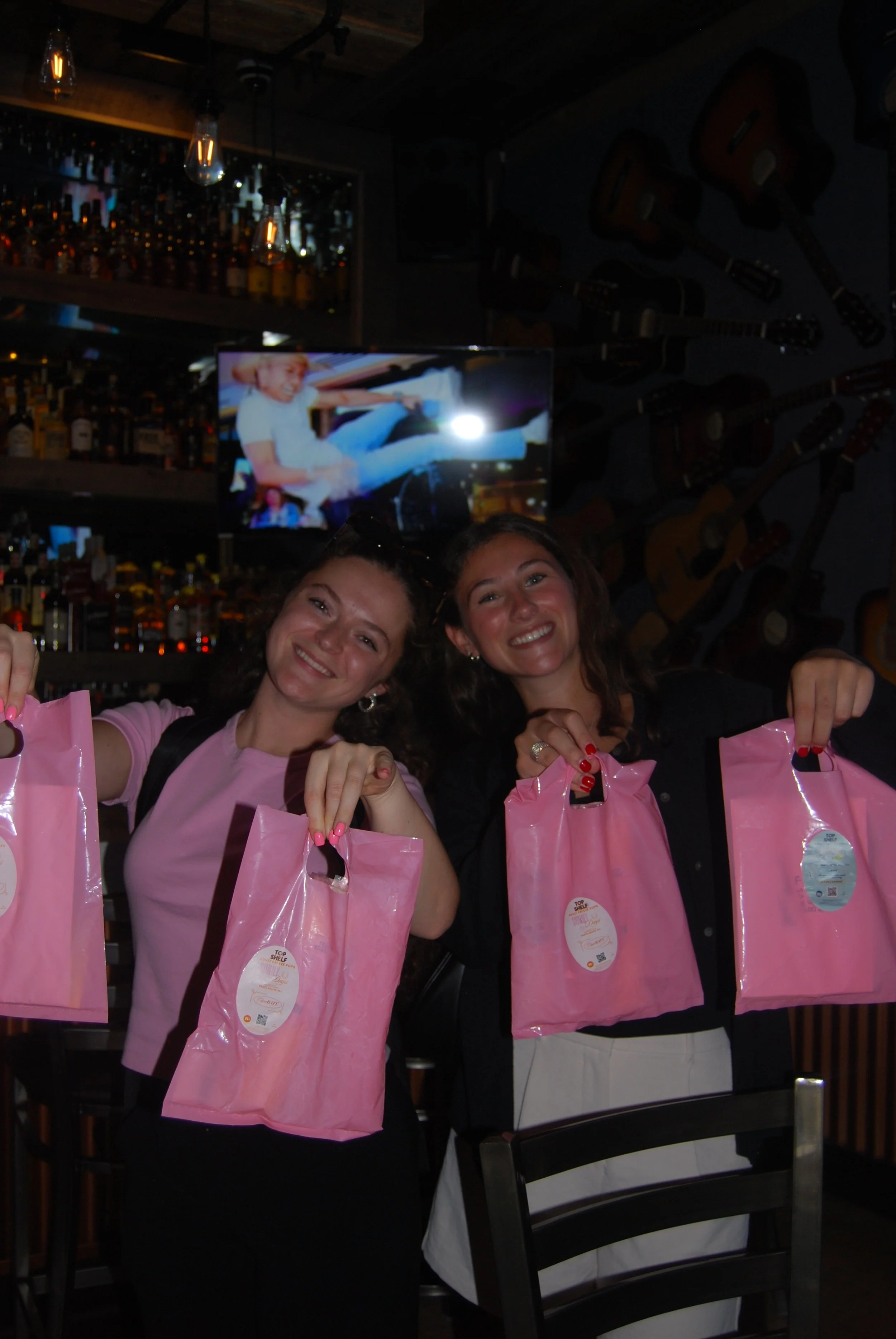 Two women smiling and holding pink bags inside a bar or restaurant with guitars on the wall and a television screen in the background.