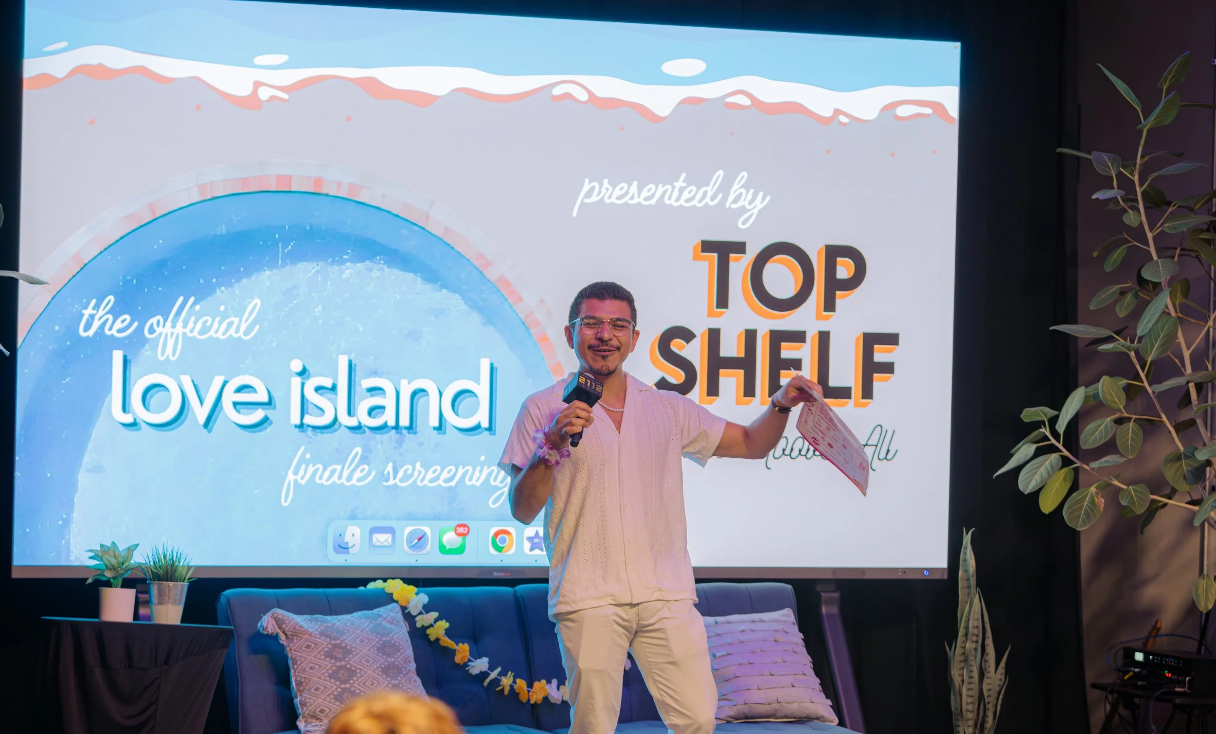A man holding a microphone and a paper, standing in front of a large screen displaying the words "the official love island finale screening" and "presented by TOP SHELF." He is smiling and wearing glasses and a short-sleeved, light-colored shirt. There are cushions and potted plants on a sofa and around the area.
