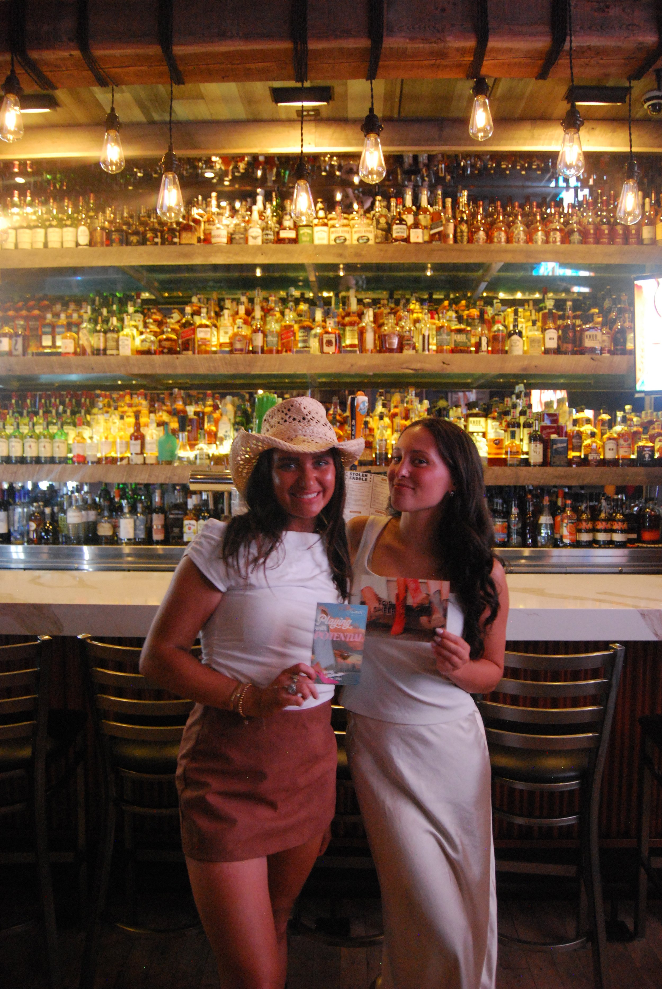Two women smiling and standing in front of a bar with numerous bottles of alcohol, one wearing a cowboy hat and the other holding a brochure.