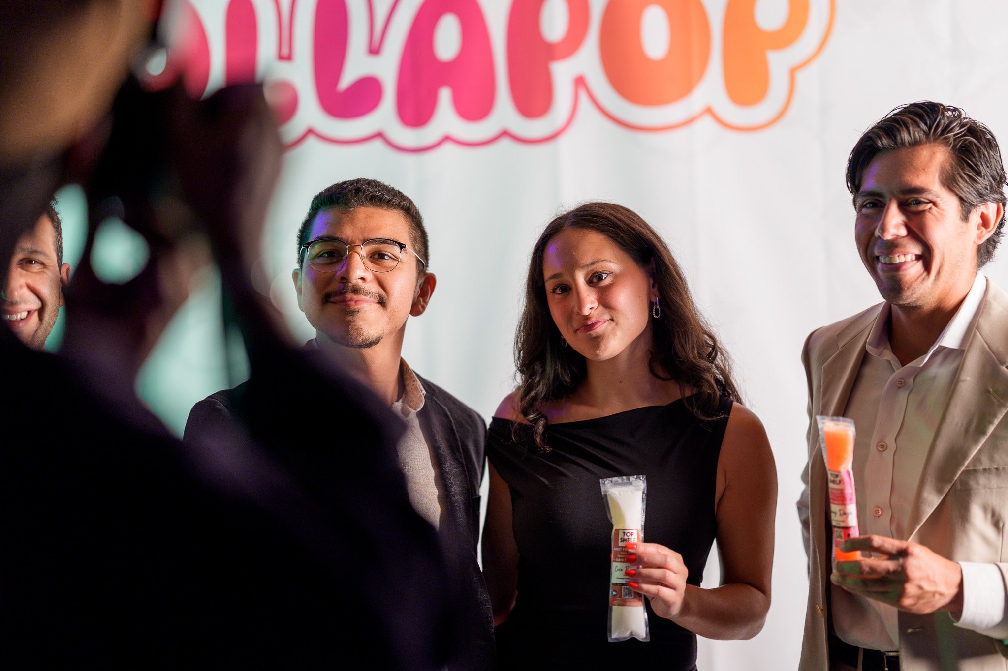 Group of five people at a celebration, with a large 'HAPPY' sign in the background. They are smiling, dressed in semi-formal attire, and holding various snacks or treats.
