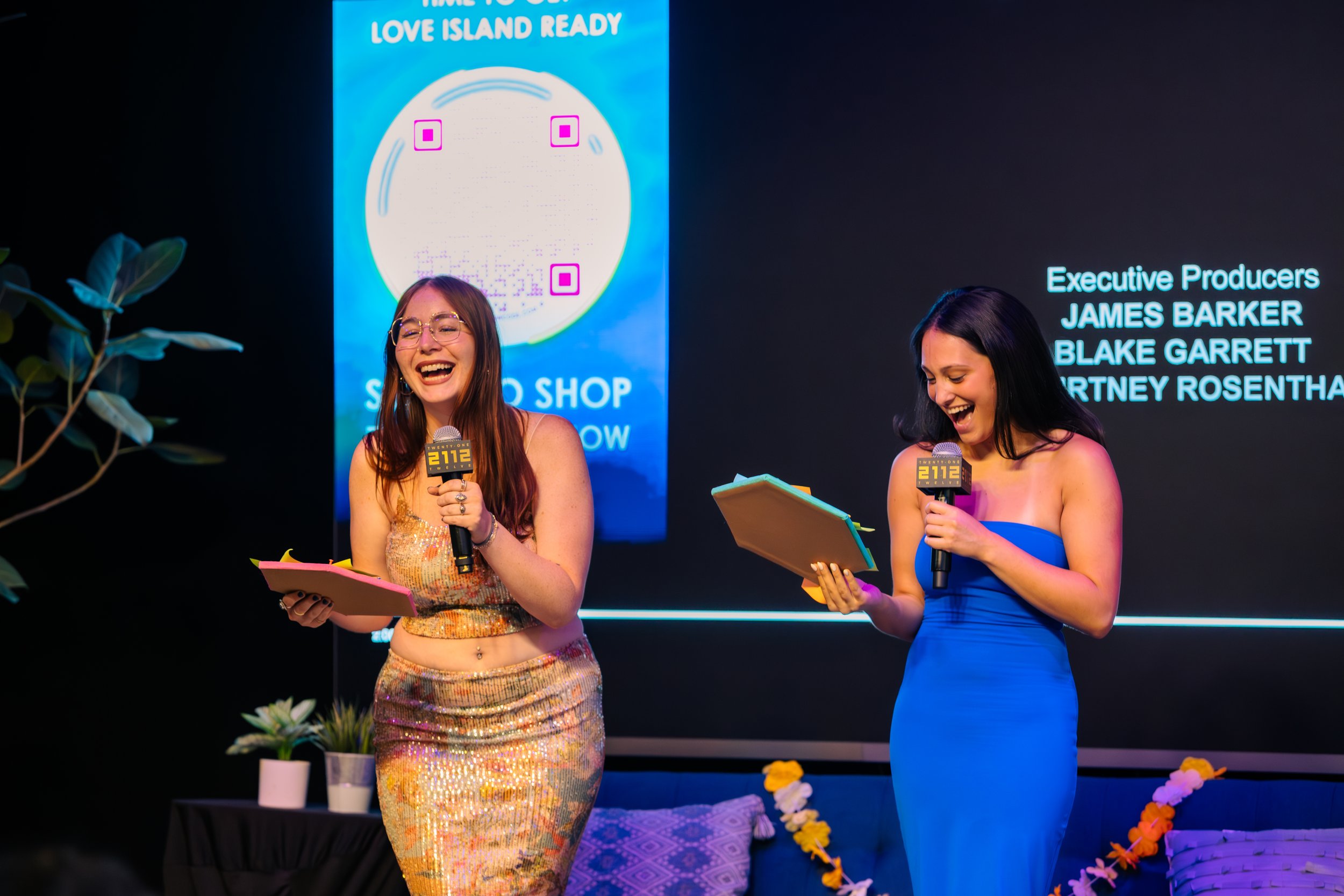 Two women on stage, laughing and holding microphones and clipboards, with a large screen behind them showing a rainbow QR code and credits for executive producers. One woman is wearing a shiny, gold, sequined dress, and the other is in a bright blue strapless dress.