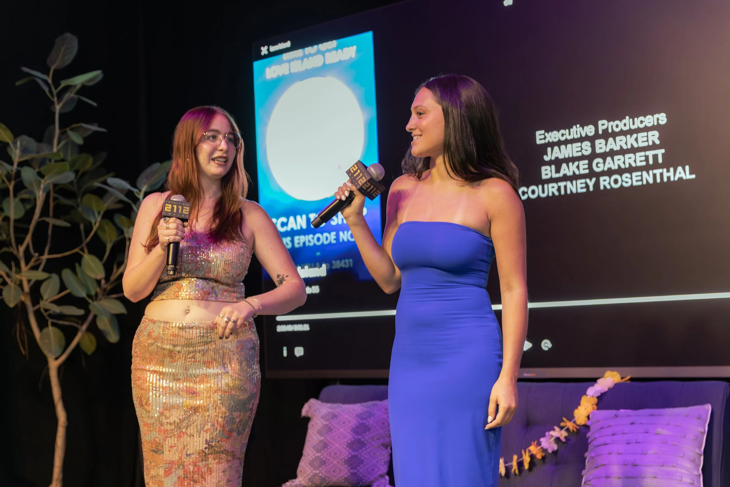 Two women are on a stage with a black background and a screen showing credits. One woman is in a sequined crop top and matching high-waisted skirt, holding a microphone. The other woman is in a strapless, form-fitting blue dress, also holding a microphone. The scene appears to be part of an interview or presentation.