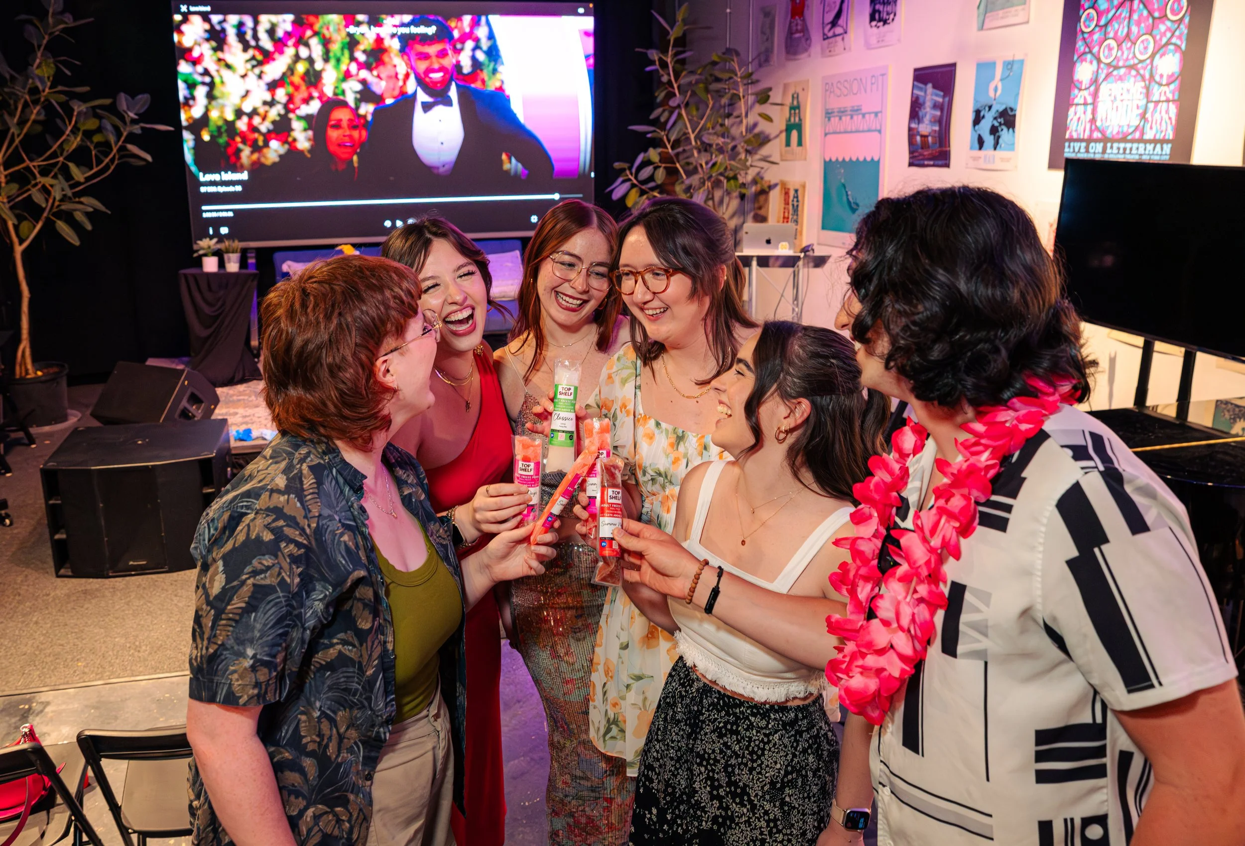 Group of seven women celebrating together, laughing, and holding skincare or beauty items at an indoor event with a backdrop of posters and a large screen showing a man in a tuxedo.