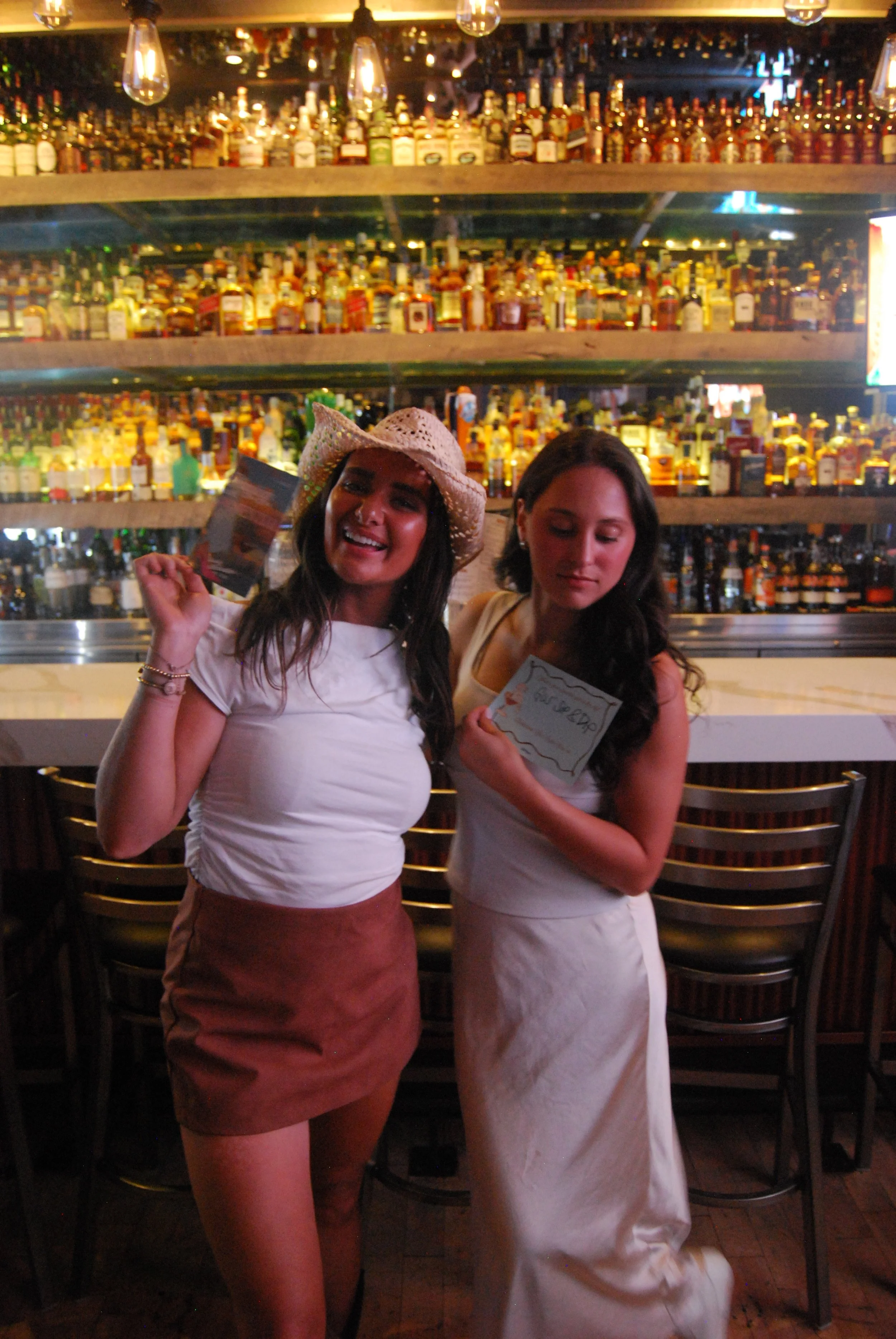 Two women standing at a bar in a lively setting with shelves filled with various liquor bottles in the background. One woman is wearing a white t-shirt, a brown skirt, and a straw hat, smiling and holding a small card. The other woman is dressed in a white dress, holding a notepad, with a neutral expression. The bar area is decorated with hanging light bulbs.