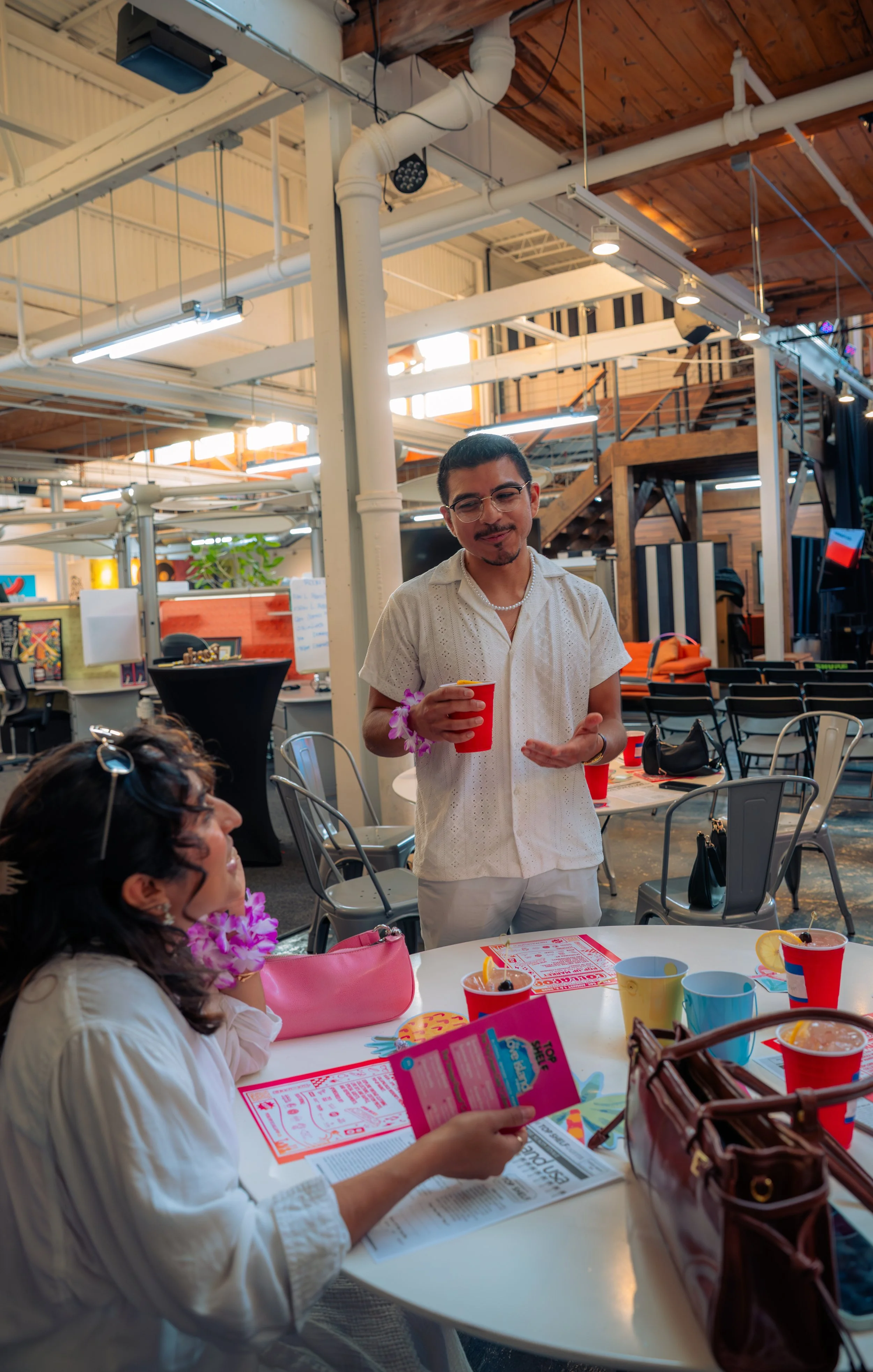 A man and woman celebrating at a party in a vibrant, open-concept indoor space with wooden accents and industrial ceiling pipes. The woman is seated, holding a menu and enjoying a drink, while the man stands nearby holding a cup and engaging in conversation.