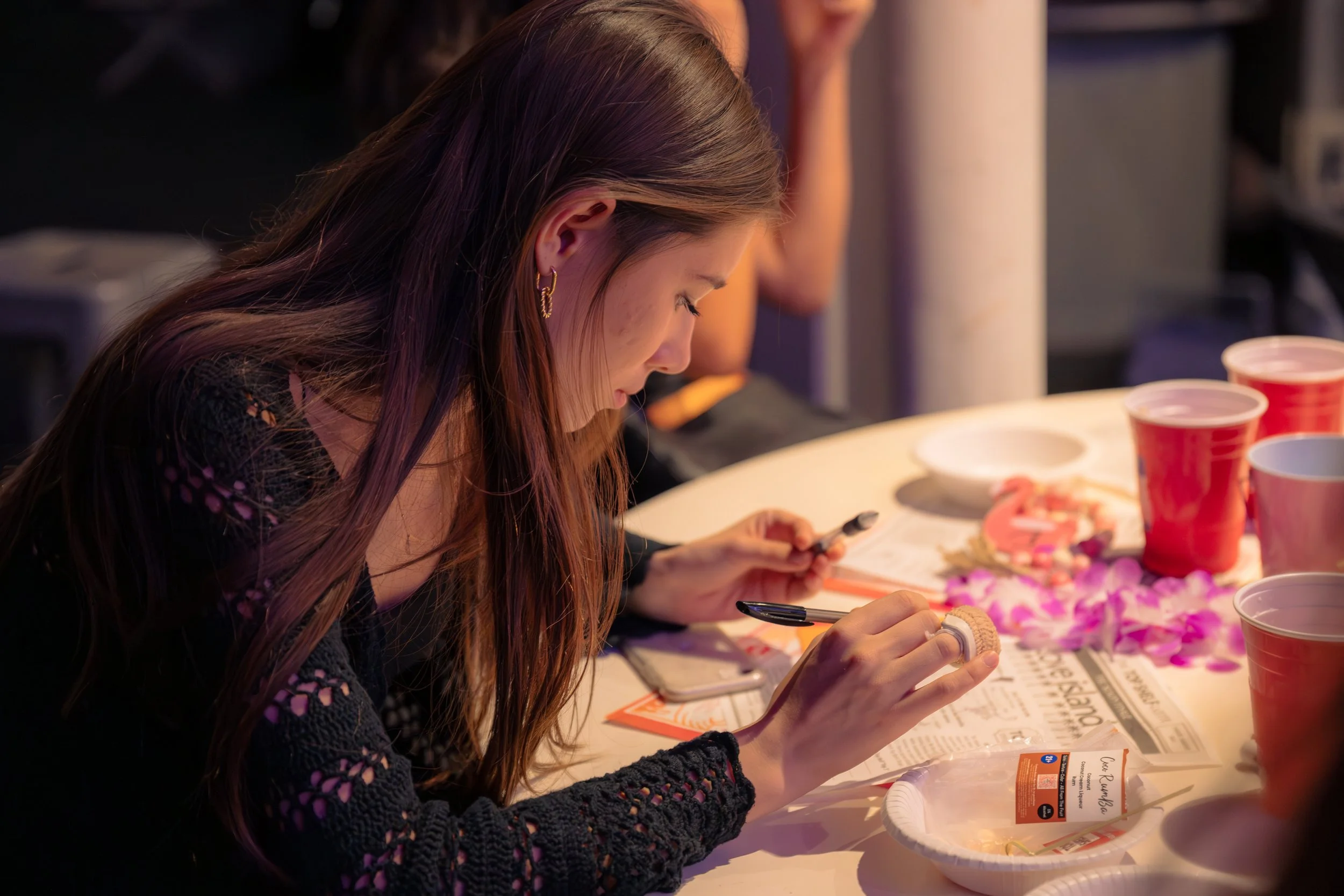 A woman with long brown hair, wearing earrings and a black lace top, is sitting at a table with several red cups, plates, and floral decorations. She is focused on taking a close-up photo of her food with her phone.
