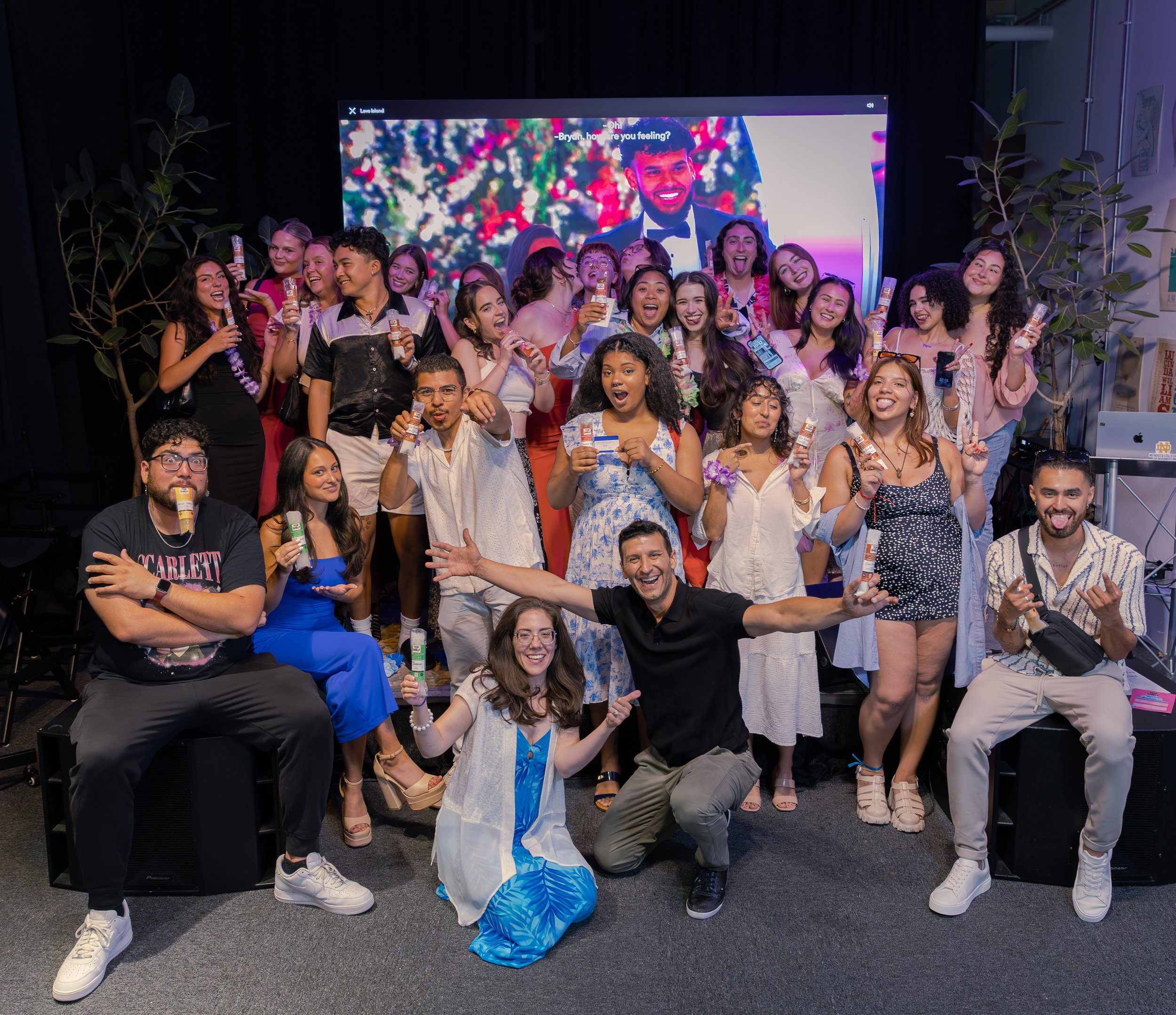 Group of diverse young adults celebrating at a party, holding drinks, smiling, and posing for the camera with a decorated background and a digital screen displaying a picture behind them.